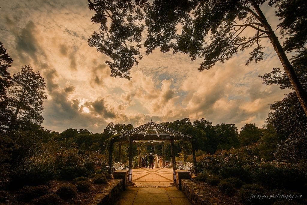 A scenic view of a gazebo in a lush garden during a dramatic sunset, perfect for a Duke Gardens wedding. The sky is filled with warm-toned clouds as a small gathering stands beneath the structure, captured expertly by a skilled photographer amid the tall trees and dense greenery.