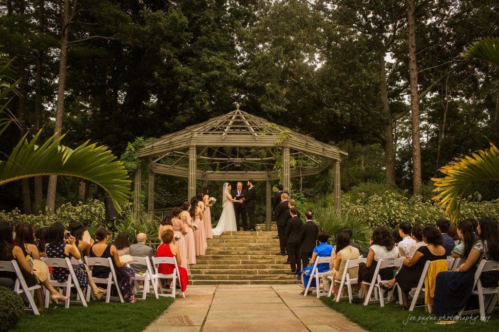 A wedding ceremony set in a lush garden with guests seated on both sides. The bride and groom stand under a wooden pergola, surrounded by greenery. Bridesmaids in pink dresses and groomsmen in black suits are near the couple.