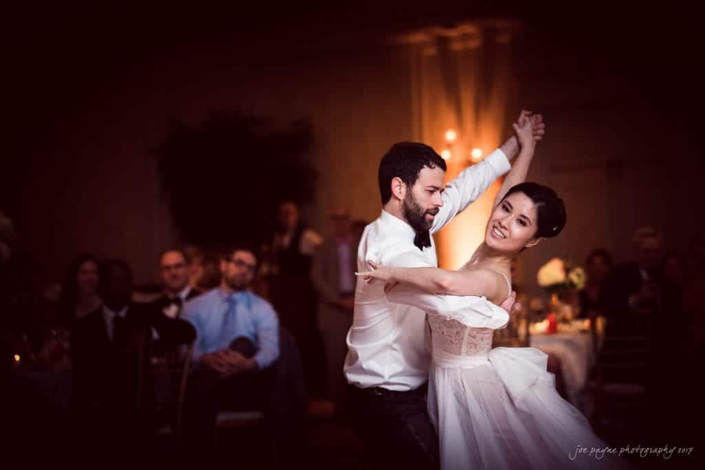 A bride and groom elegantly dance together at a wedding reception. She wears a flowing white gown, and he is in a tuxedo. The warmly lit room is filled with seated guests watching and celebrating their dance.