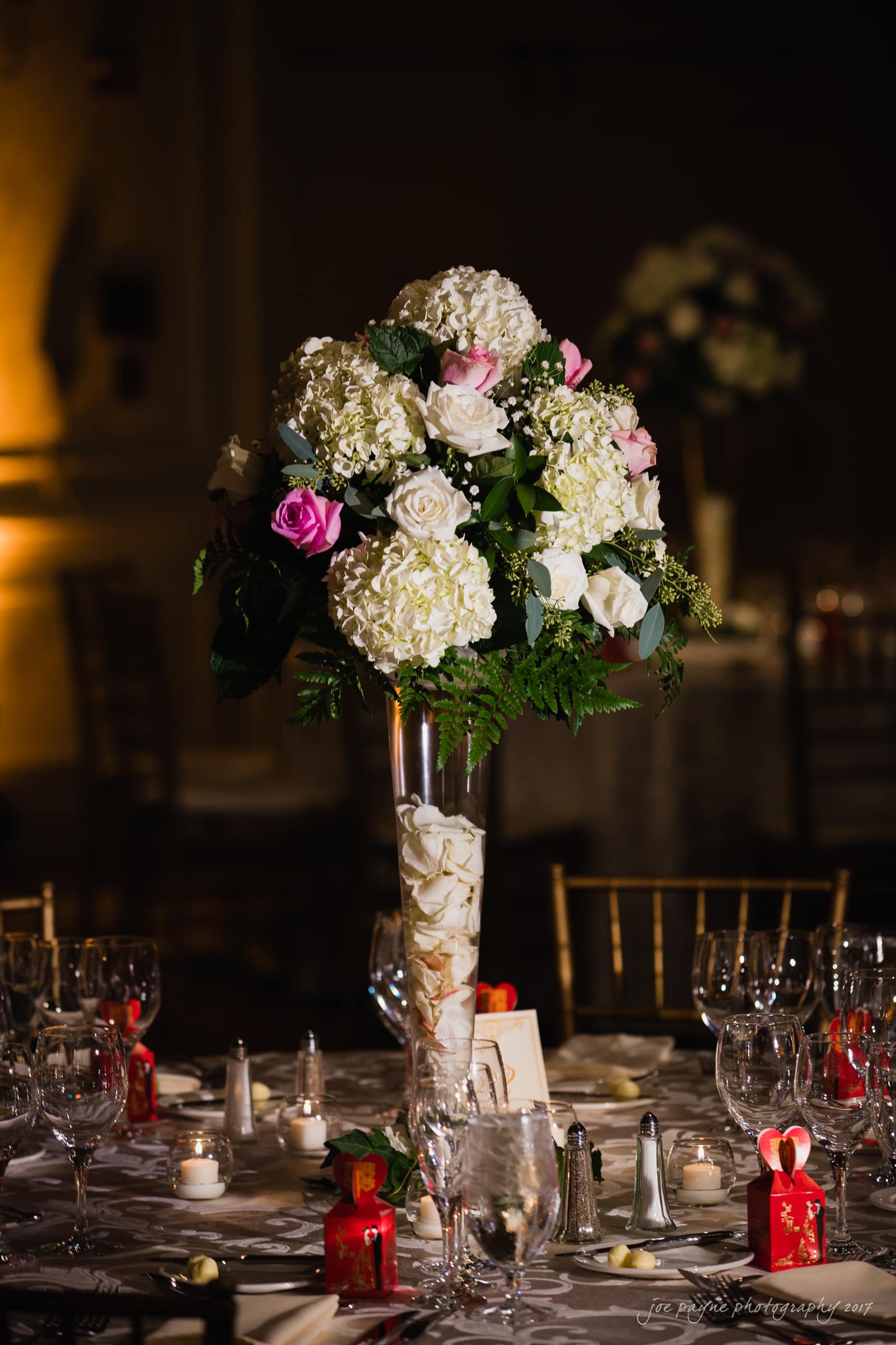 A tall floral centerpiece with white hydrangeas and pink roses in a clear vase, set on an elegantly decorated dining table with lit candles and small red gift boxes. The background is dimly lit, highlighting the arrangement.