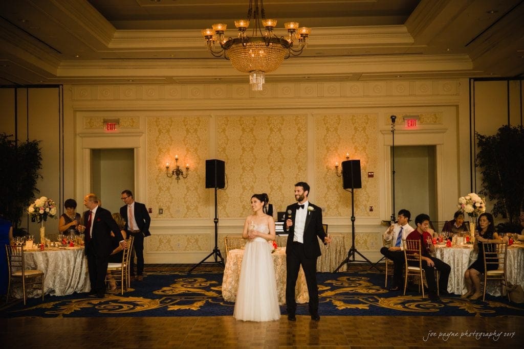A bride in a white gown and a groom in a black tuxedo stand together on a ballroom dance floor, captured beautifully by a Duke Gardens wedding photographer. Guests seated at tables are visible in the background under elegant chandeliers.
