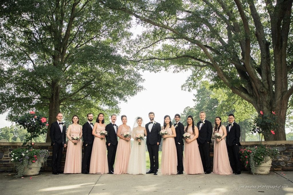A wedding party poses outdoors under trees. The bride and groom stand in the center; bridesmaids in peach dresses and groomsmen in black suits flank them. They all hold bouquets and smile for the photo.