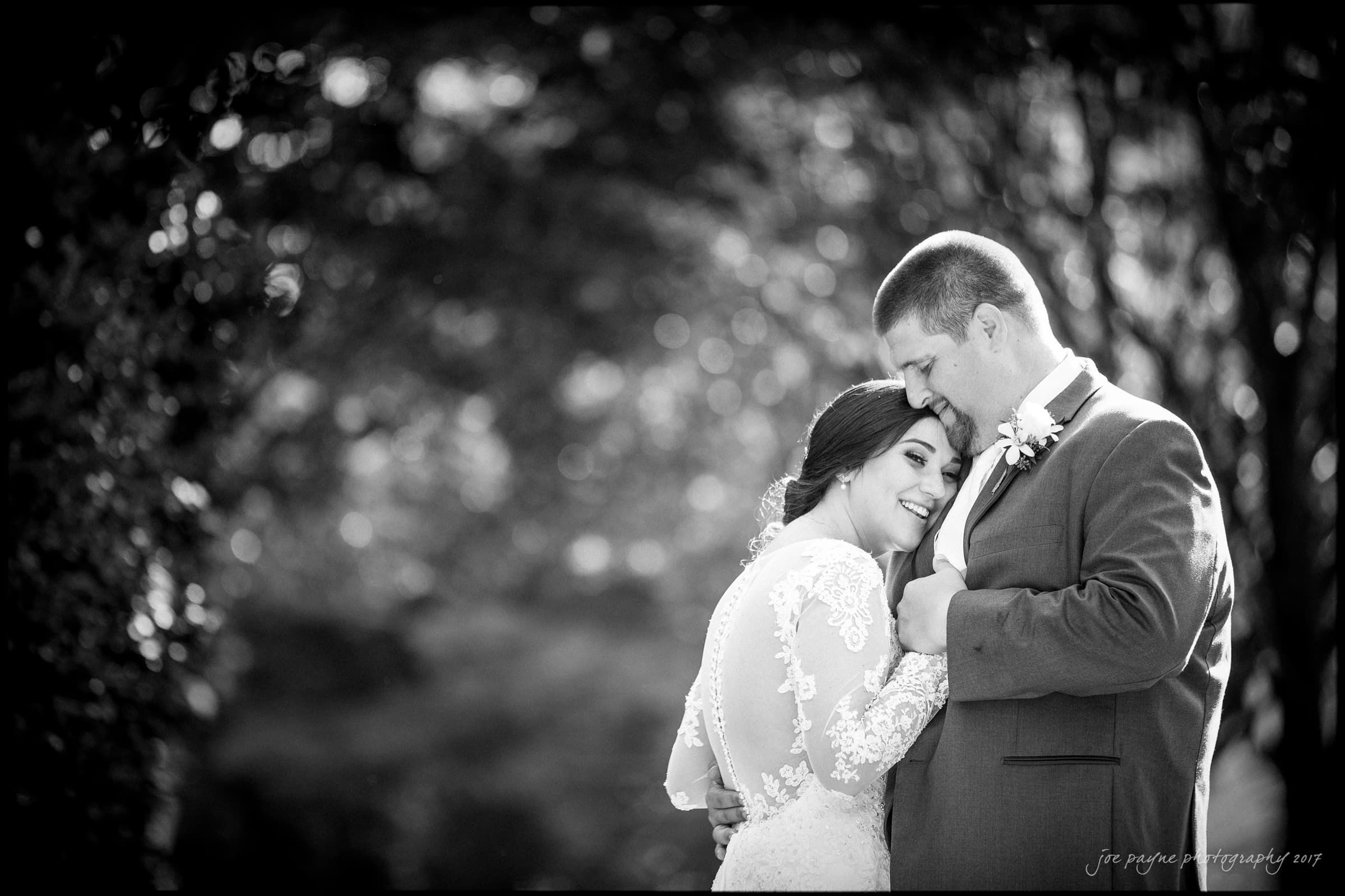 A black and white photo captures a smiling bride in a lace dress and a groom in a suit. The groom gently rests his forehead against hers, surrounded by a softly focused natural background.