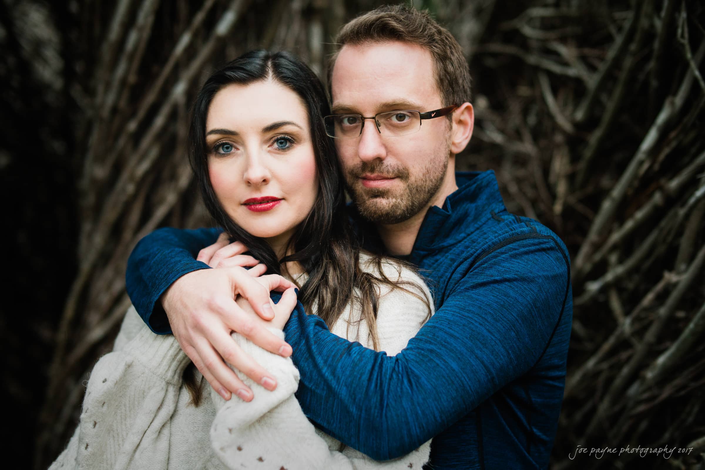 A man and woman stand closely together, with the mans arms around her shoulders. They both have serious expressions. The background features intertwining branches, adding a natural touch to the scene.