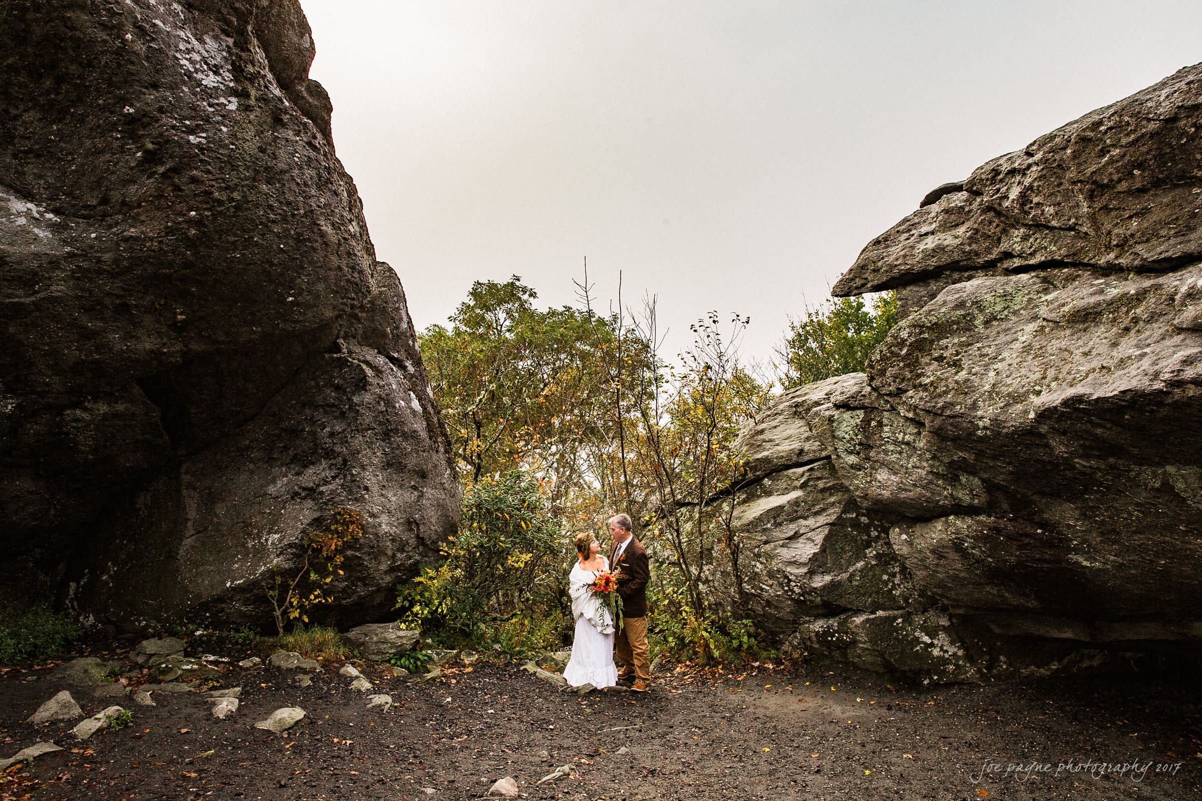 A couple in wedding attire stand between two large rock formations, surrounded by greenery. The bride holds a bouquet, and the groom is in a suit. The scene has a natural, outdoor setting with a cloudy sky.
