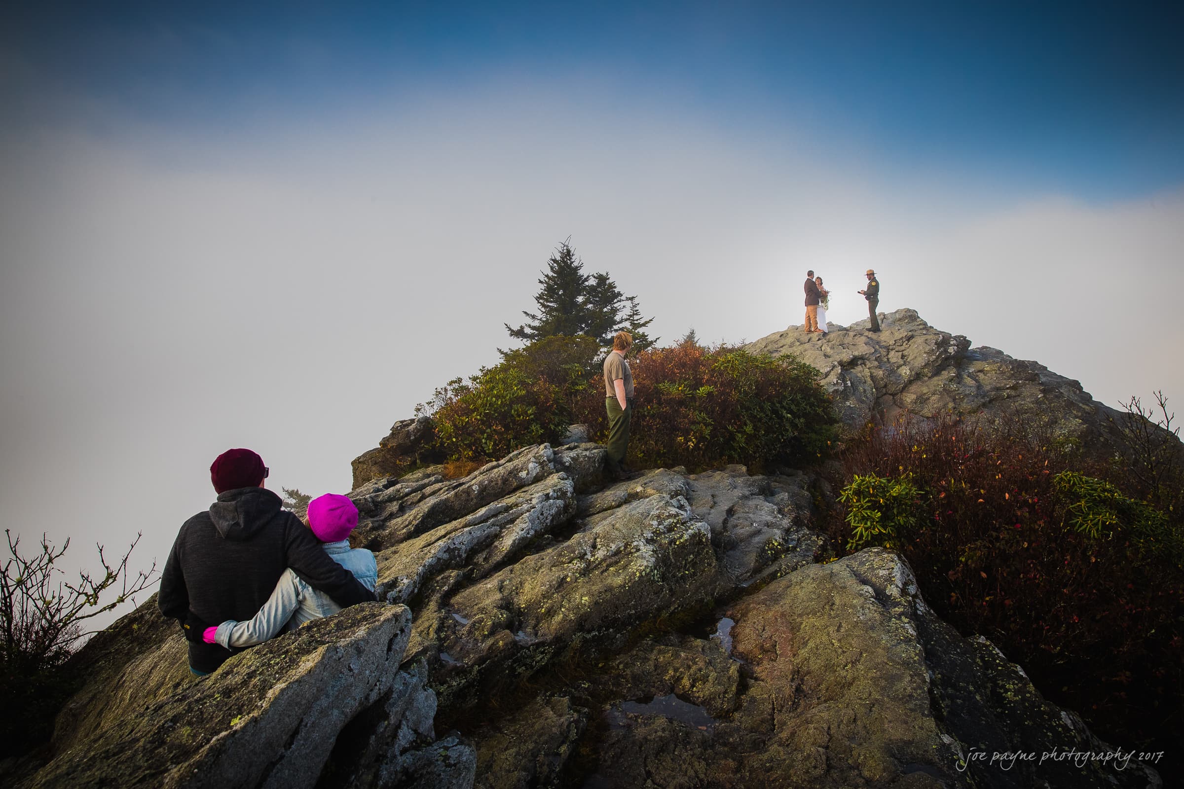 A couple with a child sits on rocky terrain, wearing warm clothes. Higher up, two people stand on the summit beneath a bright blue sky, surrounded by fog. Small trees and shrubs dot the rocky landscape.