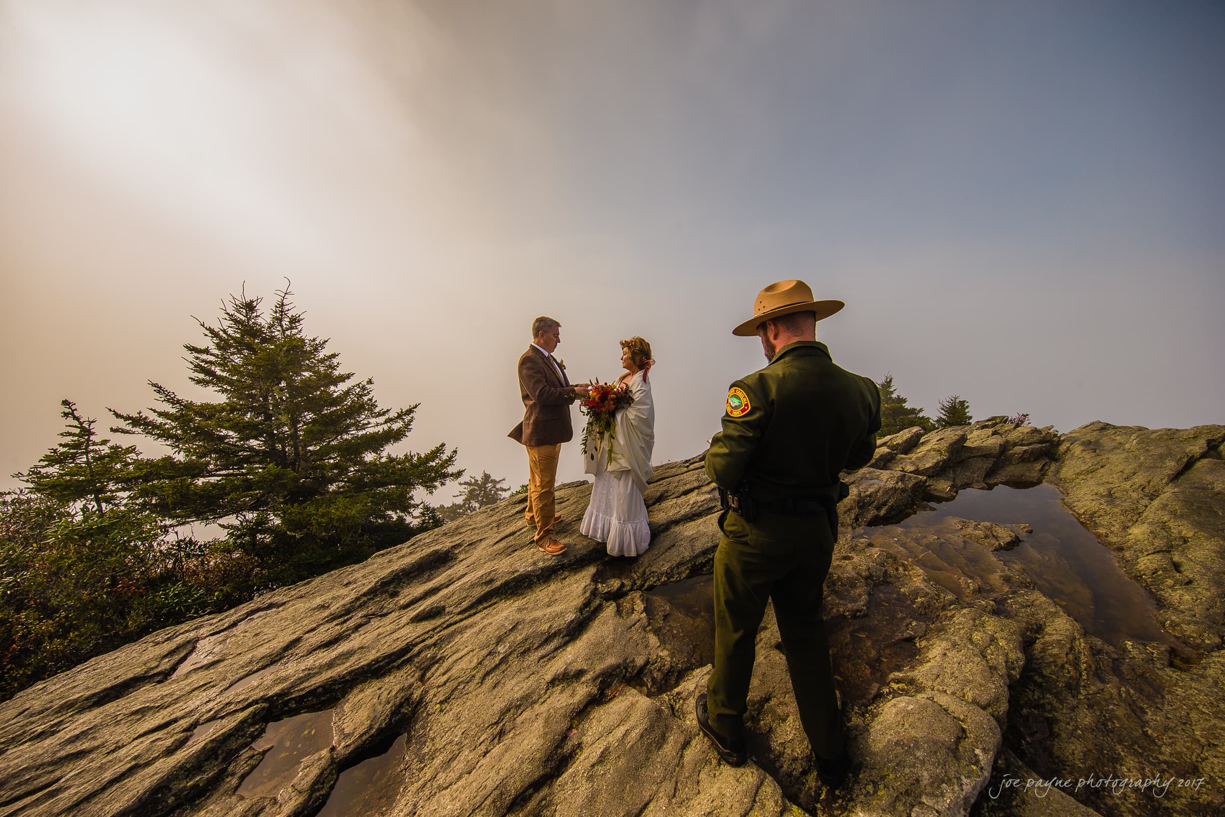 A couple stands on a rocky cliff exchanging vows during their wedding ceremony with an officiant in a park ranger uniform. The background is misty, and there are evergreen trees nearby.