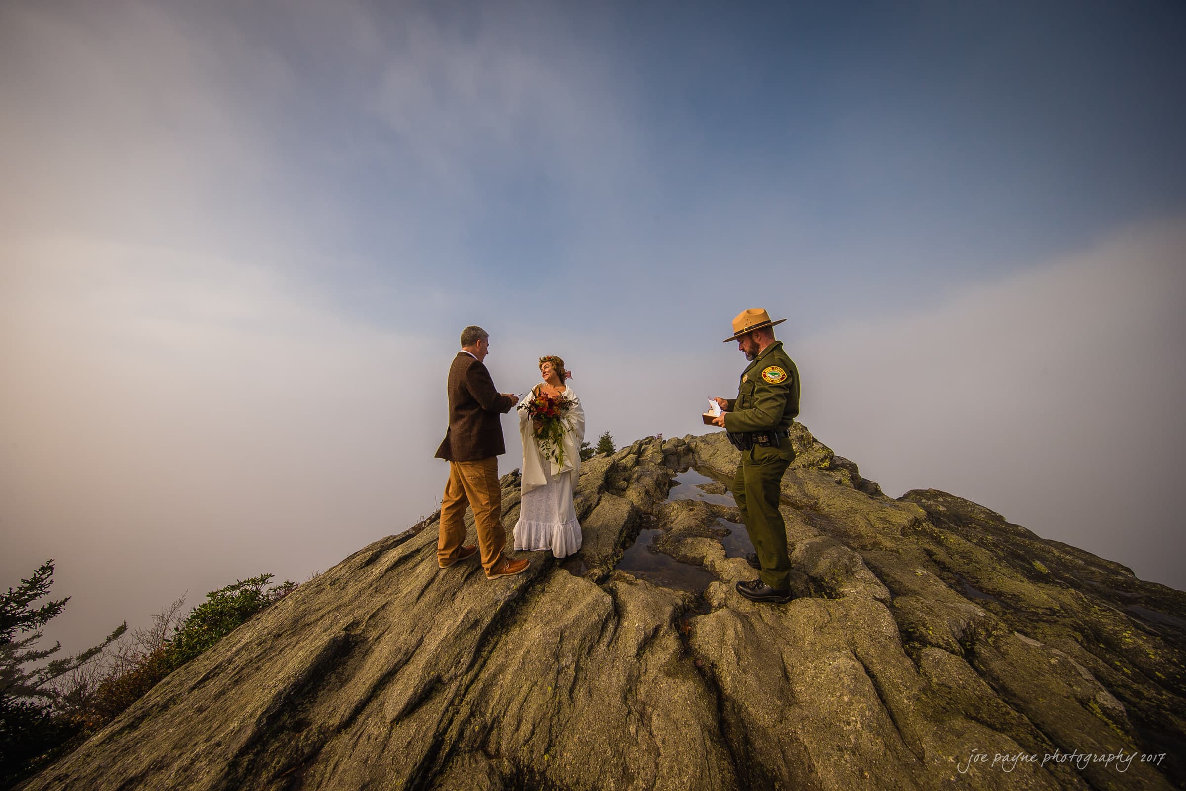 A bride and groom stand on a rocky summit during a foggy outdoor wedding ceremony, accompanied by a park ranger officiating. The landscape is rugged, and the sky is partly cloudy, adding a dramatic backdrop to the intimate setting.