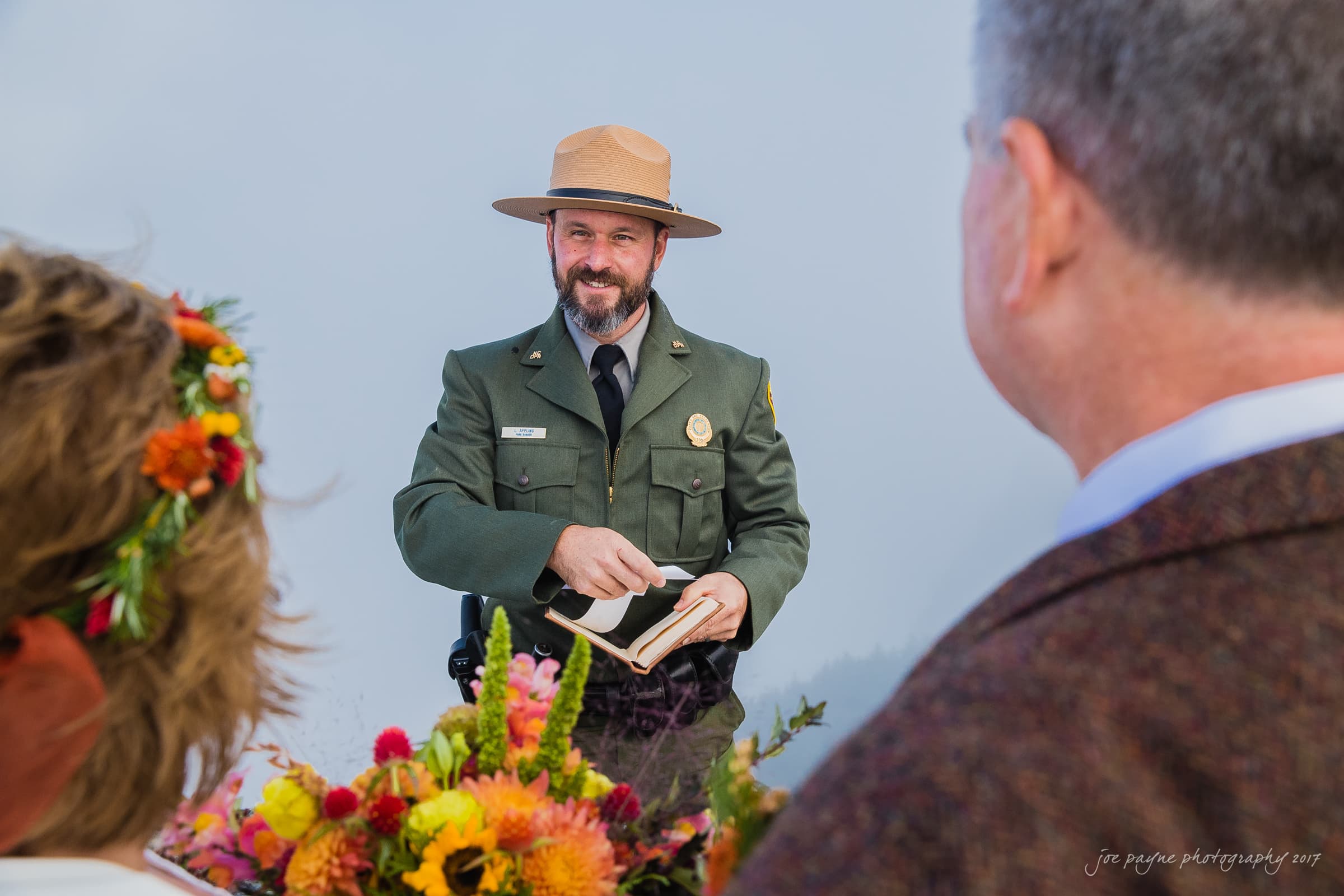 A park ranger officiates a wedding ceremony outdoors. The ranger is wearing a green uniform and brown hat while holding a book. The couple, with their backs turned, are adorned with colorful flowers. A misty landscape is visible in the background.