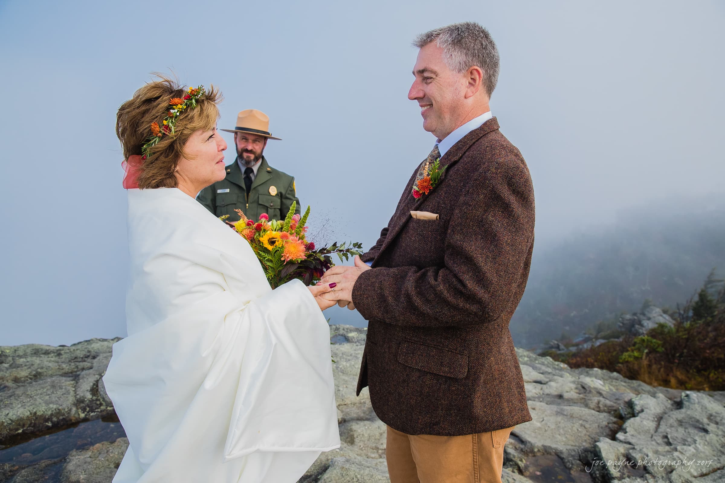 A couple holds hands during an outdoor wedding ceremony on a foggy day. The bride is wrapped in a white shawl, holding a colorful bouquet, and the groom is in a brown suit. A park ranger stands behind them officiating. The setting is rocky and misty.