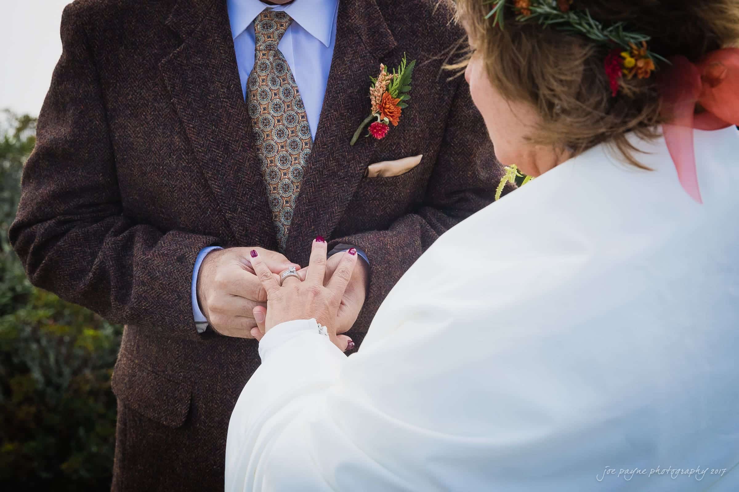 A couple is exchanging rings outdoors. The person on the right is placing a ring on the others finger. Both are dressed in formal attire, with the left wearing a suit and tie, and the right adorned with a floral headpiece.