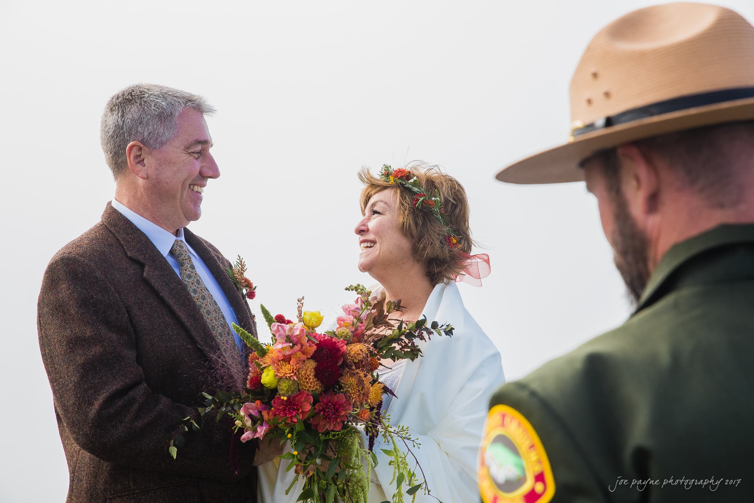 A couple, dressed in a suit and a white shawl with a bouquet, smile warmly at each other during a wedding ceremony. A park ranger stands beside them, wearing a uniform and hat. The sky is overcast, adding a soft backdrop to the joyful scene.