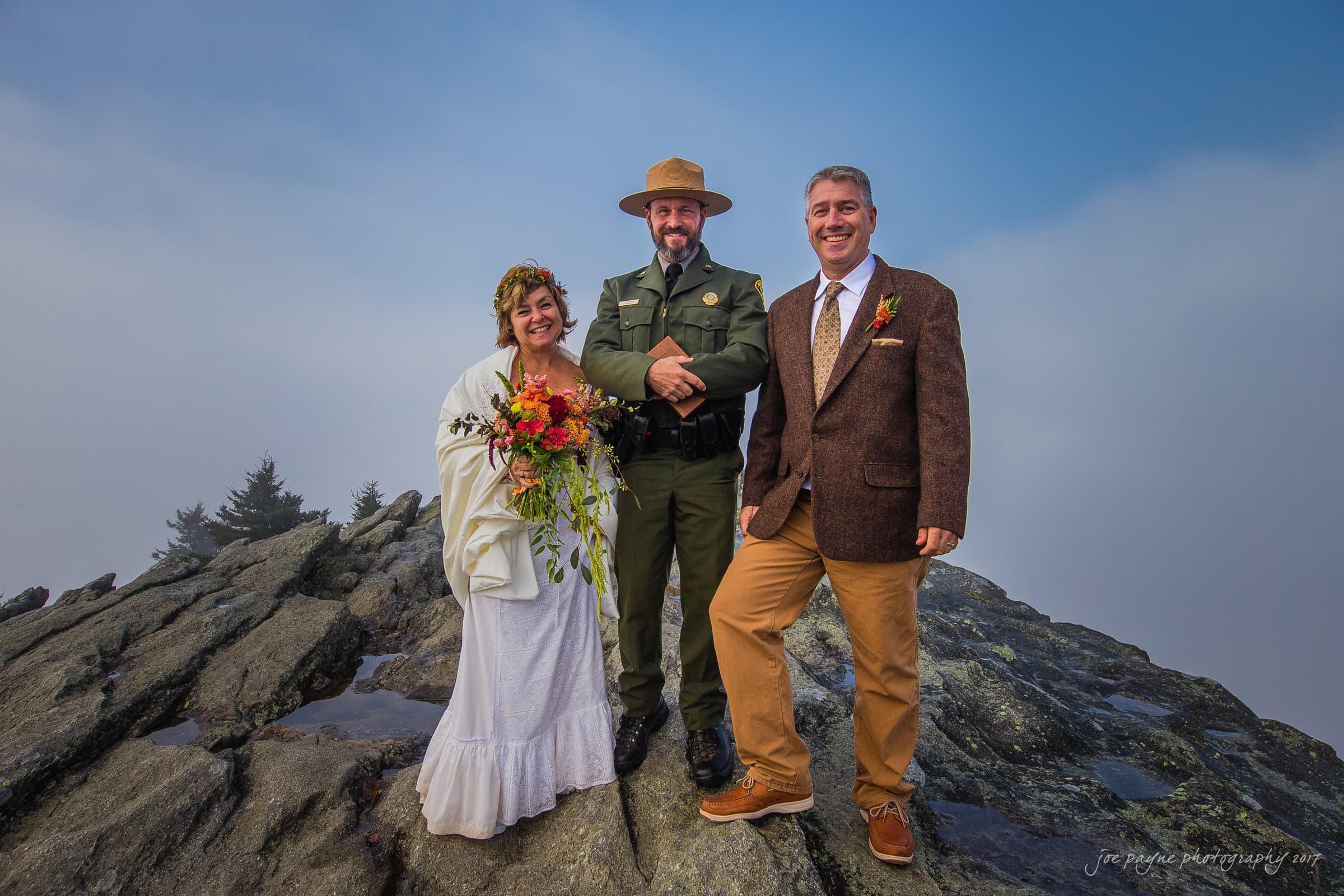 A couple stands on a rocky mountain peak under a cloudy sky during their wedding ceremony. The bride holds a bouquet and wears a white dress, while the groom wears a brown suit. A park ranger stands between them, smiling.