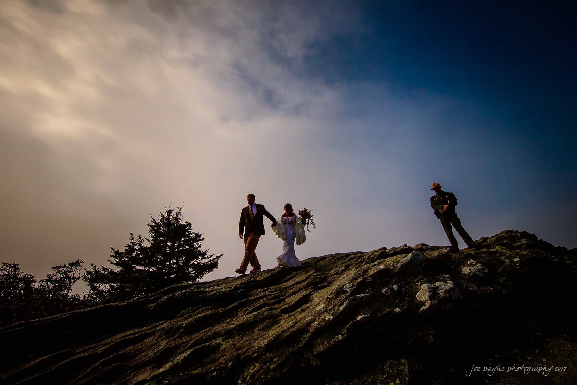 A couple in wedding attire walks hand in hand across a rocky hilltop, silhouetted against a dramatic sky. A person stands nearby, observing the scene. A lone tree and cloudy background enhance the moody atmosphere.