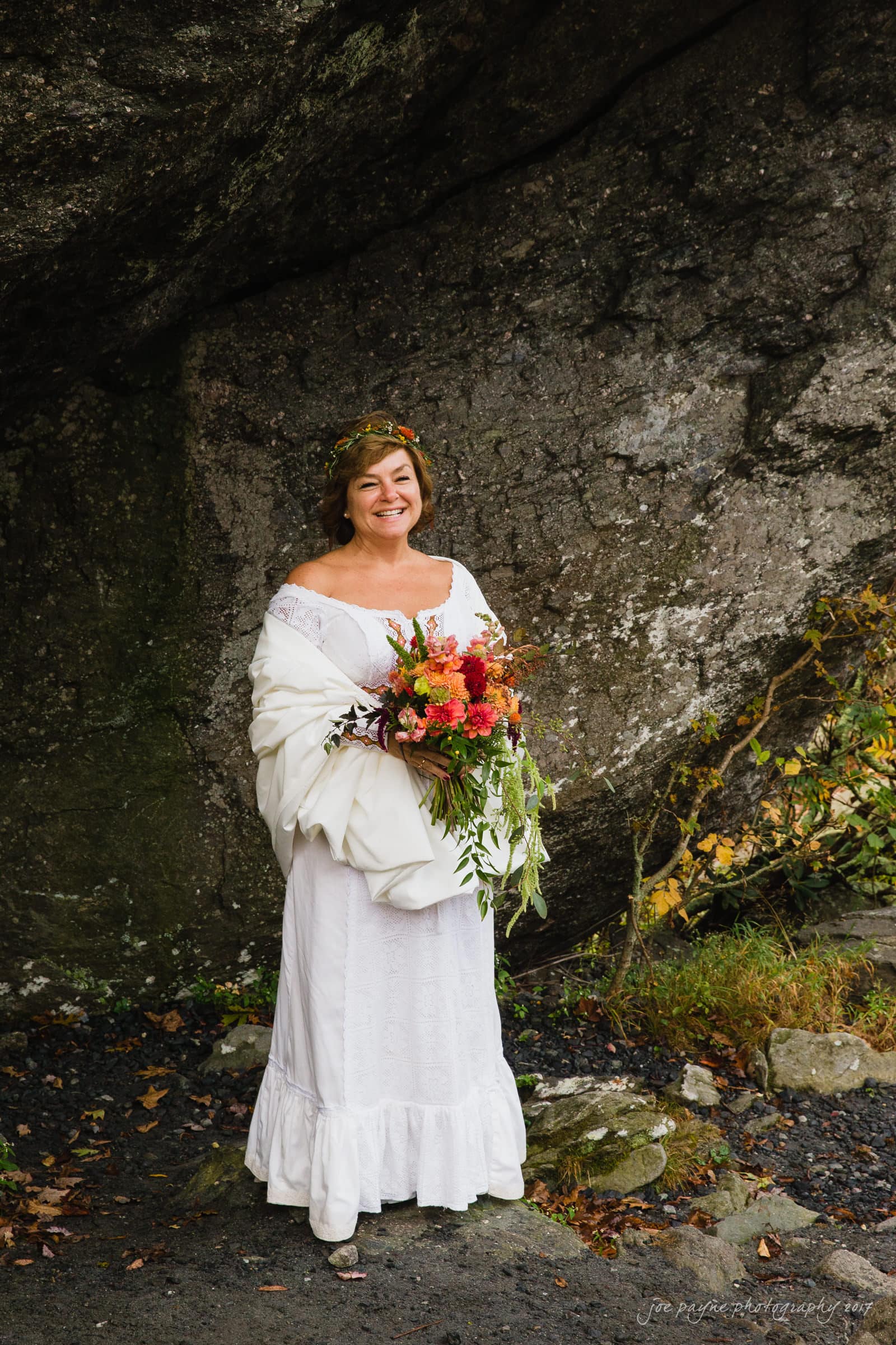A bride in a white dress, with a shawl draped over her shoulders, stands smiling and holding a vibrant bouquet. She is in front of a rocky backdrop with some foliage and wearing a floral headpiece.