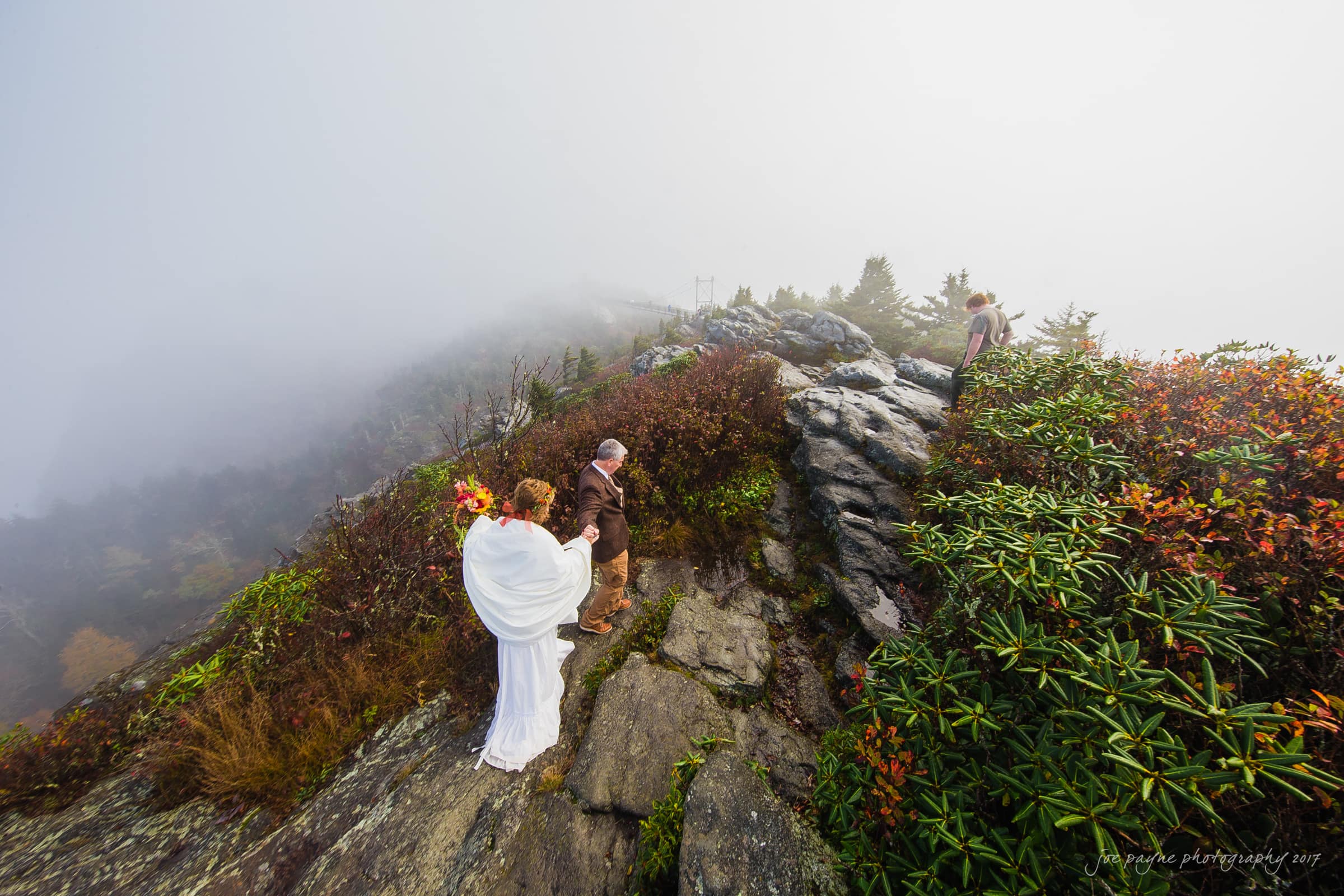 A couple stands on a rocky, foggy mountaintop exchanging vows. The bride wears a white dress and holds a bouquet, while the groom is dressed in a brown outfit. Mist surrounds them, and fall foliage adds color to the scene.