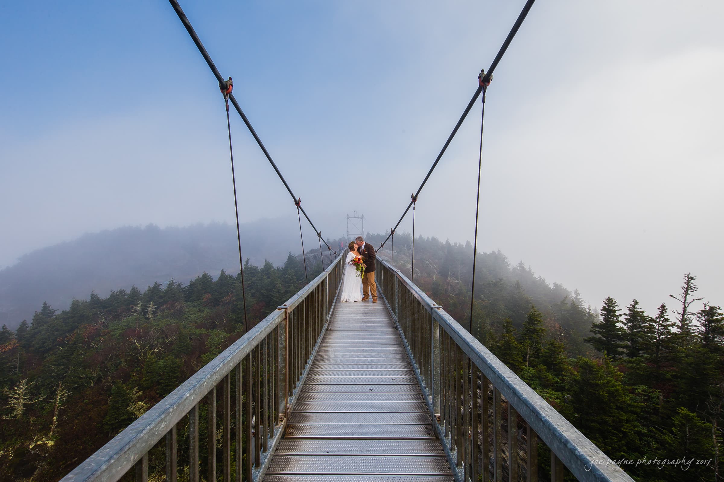 A couple stands at the center of a misty suspension bridge surrounded by forested mountains. The sky is foggy, and the perspective of the bridge emphasizes the expansive, scenic view.