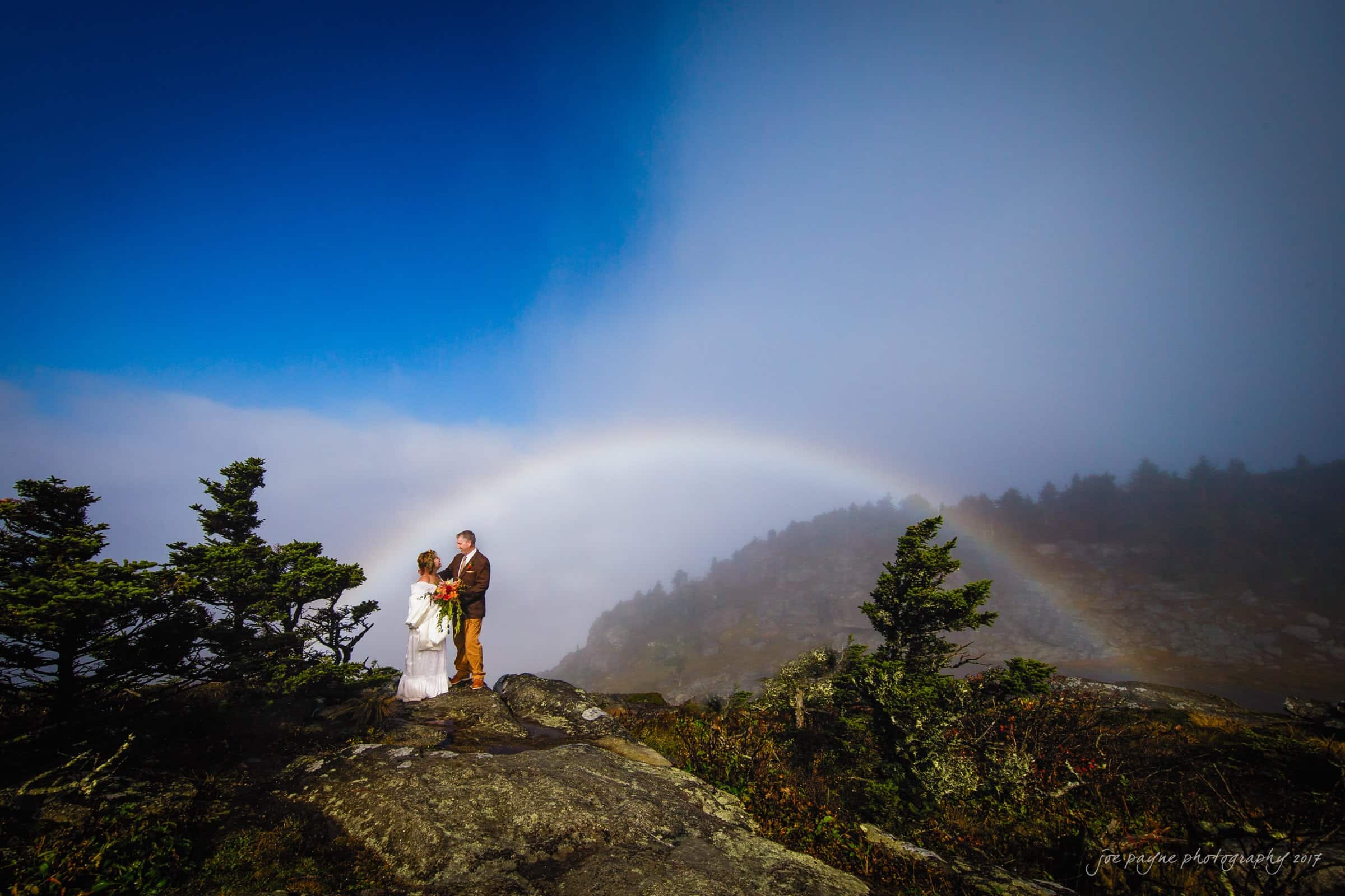 A bride and groom pose on a rocky mountain ledge surrounded by mist and greenery. A faint rainbow arches across the cloudy sky, adding a serene touch to the vibrant setting.
