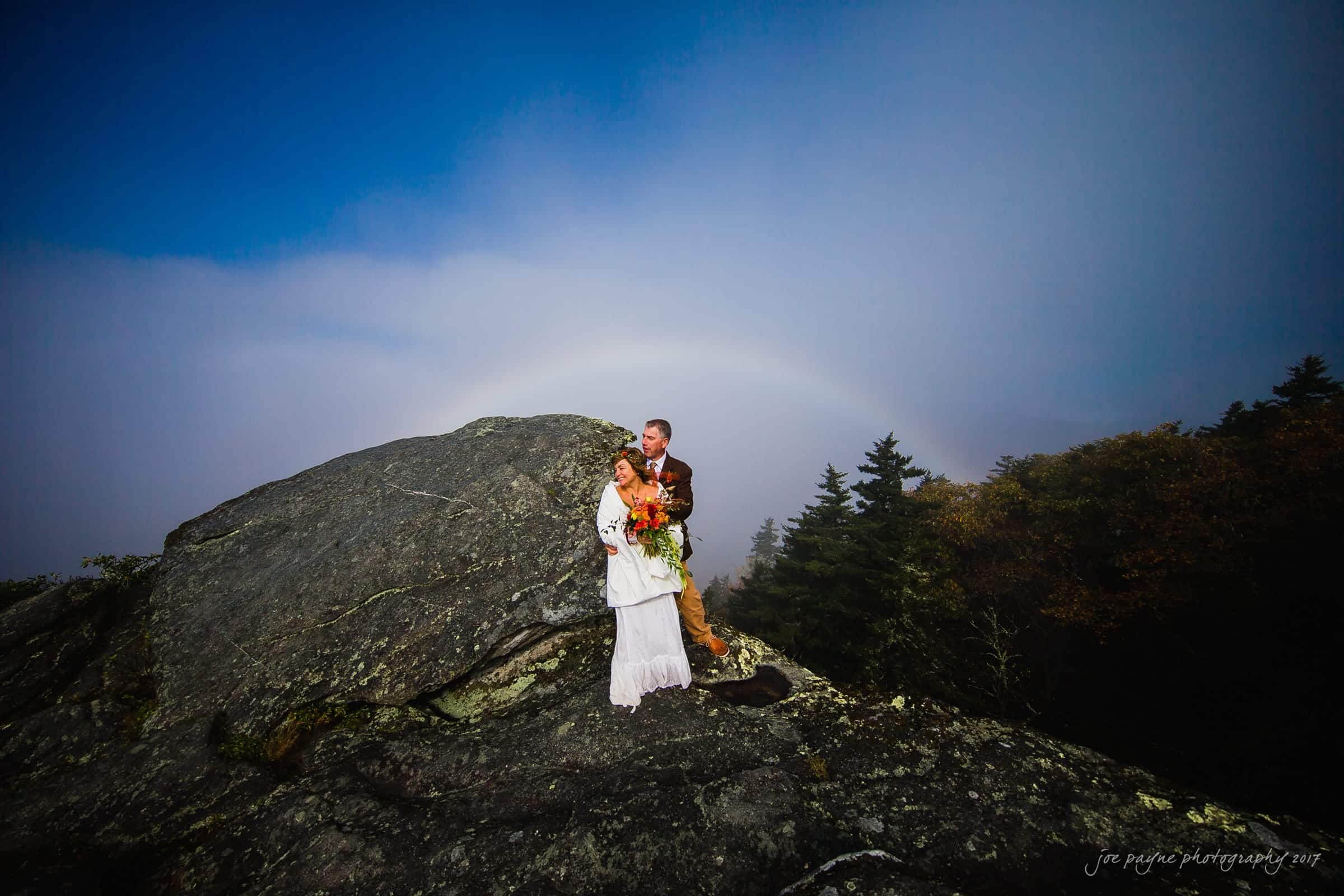 A couple stands on a rocky hilltop under a vibrant rainbow. The woman in a white dress holds a bouquet of orange flowers, while the man in a brown jacket has his arm around her. The sky is overcast, and trees are visible in the background.