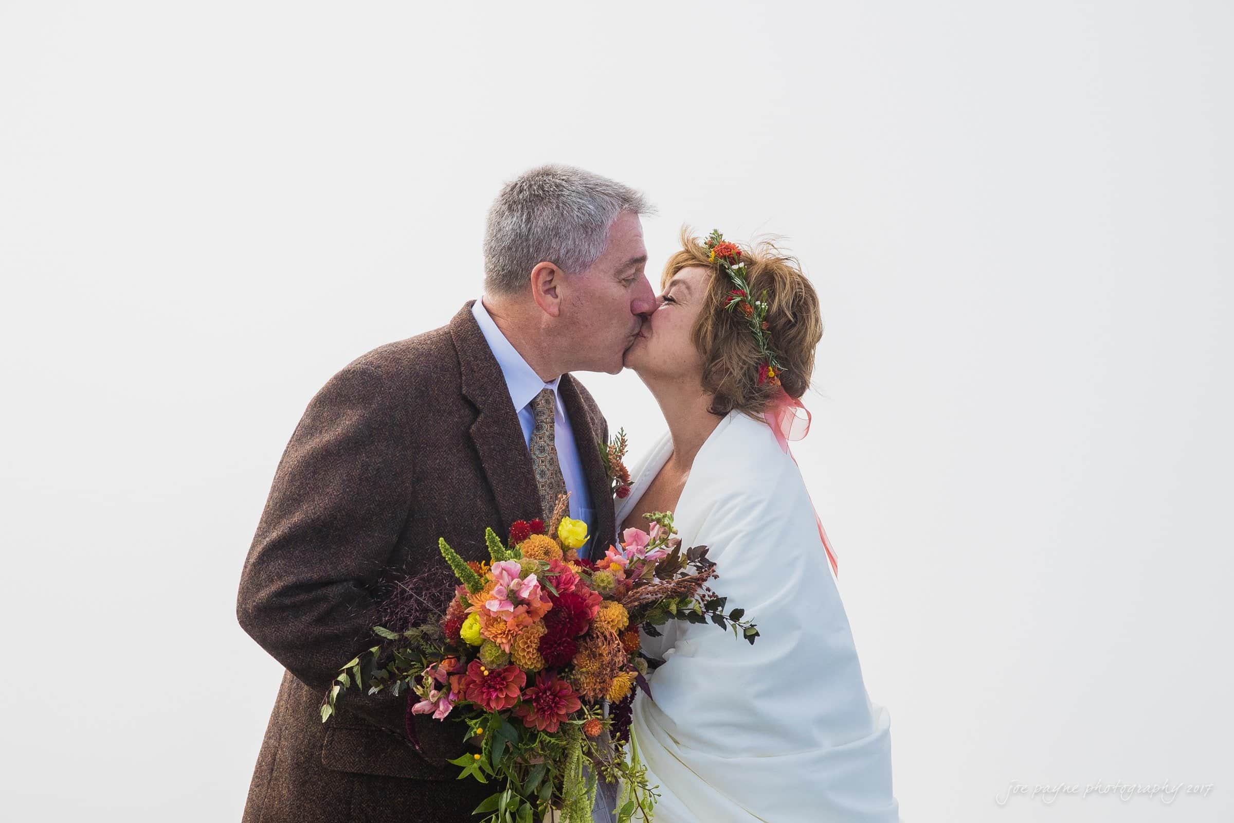 A couple shares a kiss, standing close together. The woman holds a vibrant bouquet of flowers, and both are dressed warmly. The background is a soft, pale color, creating an intimate and serene atmosphere.