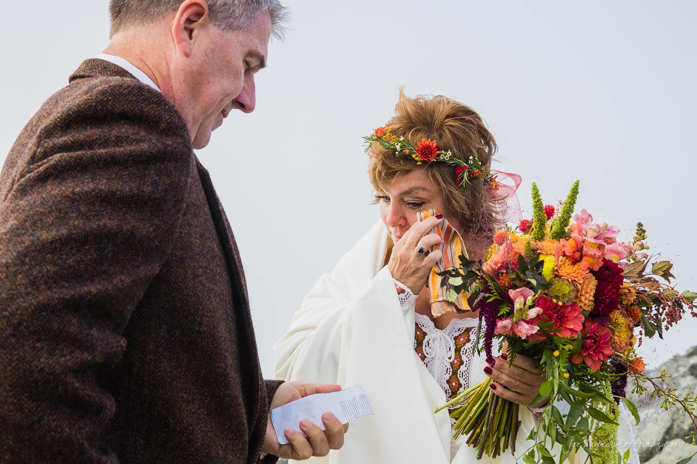 A woman wipes her tears while holding a colorful bouquet, standing next to a man in a suit who is holding a piece of paper. She wears a floral crown and a decorated shawl, creating a tender and emotional scene.