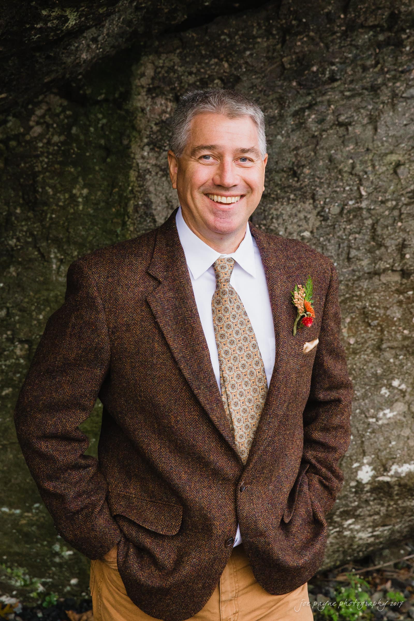 A smiling man stands outdoors in front of a rocky background. He is wearing a brown tweed blazer, a white shirt, and a patterned tie, with hands in his pockets. His blazer is accessorized with a small boutonniere.