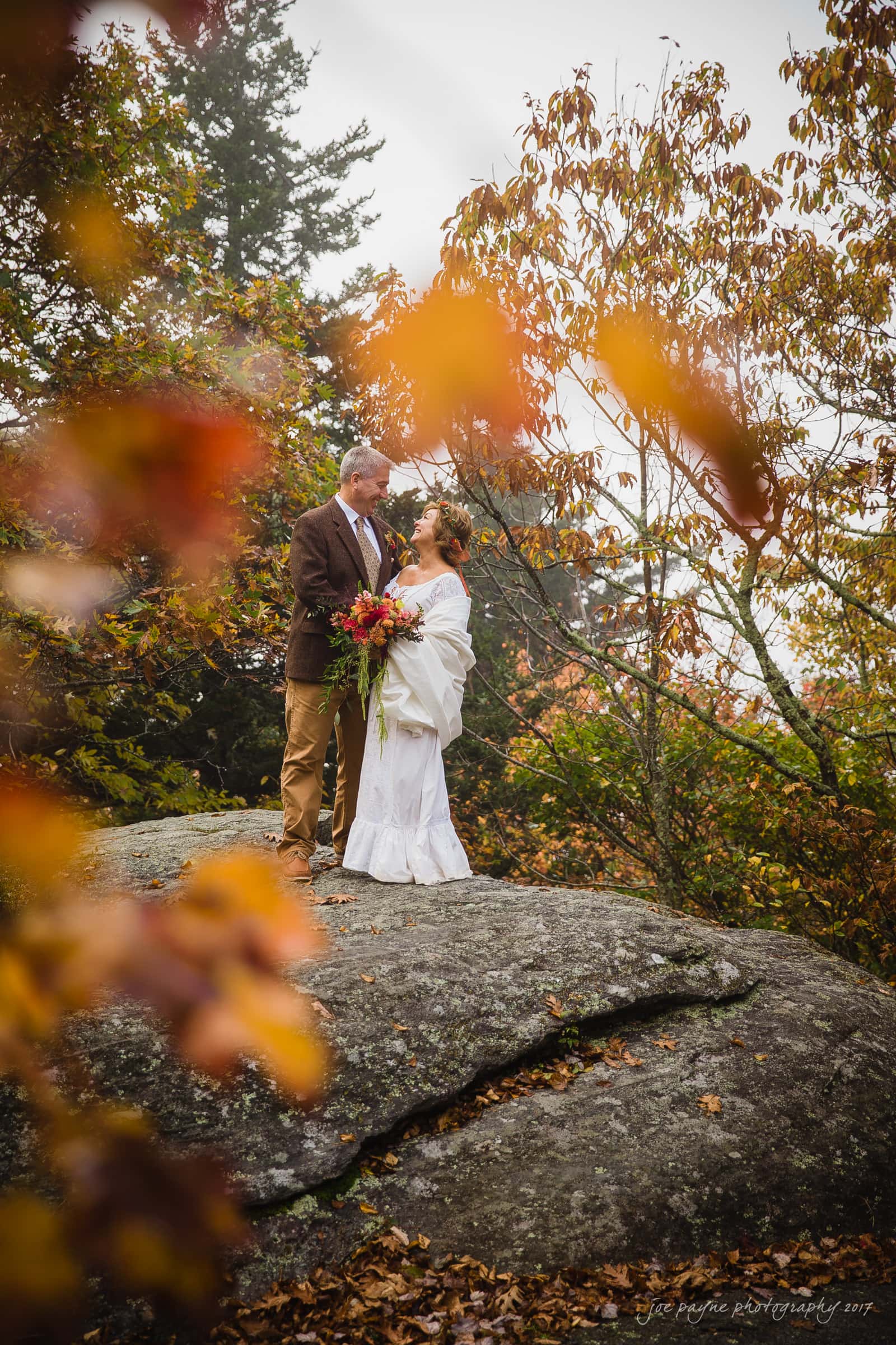 A couple in wedding attire stands on a large rock surrounded by autumn foliage. The bride holds a bouquet of red and orange flowers. They are gazing at each other amidst a scenic backdrop of trees with colorful leaves.