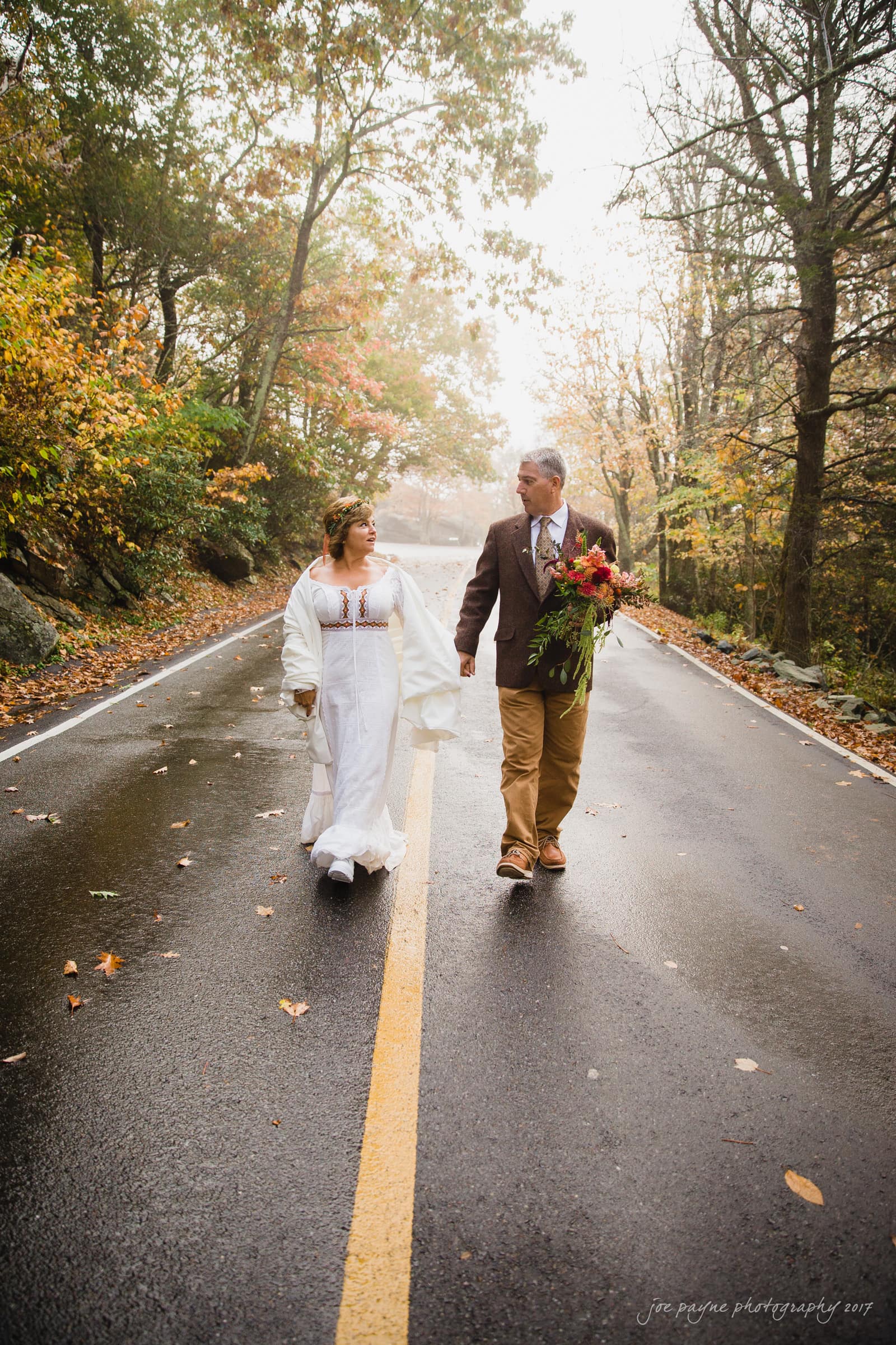A couple walks hand in hand down a misty, tree-lined road. The woman wears a white dress, and the man is in a brown jacket, holding a bouquet. Autumn leaves in shades of orange and yellow frame the scene.