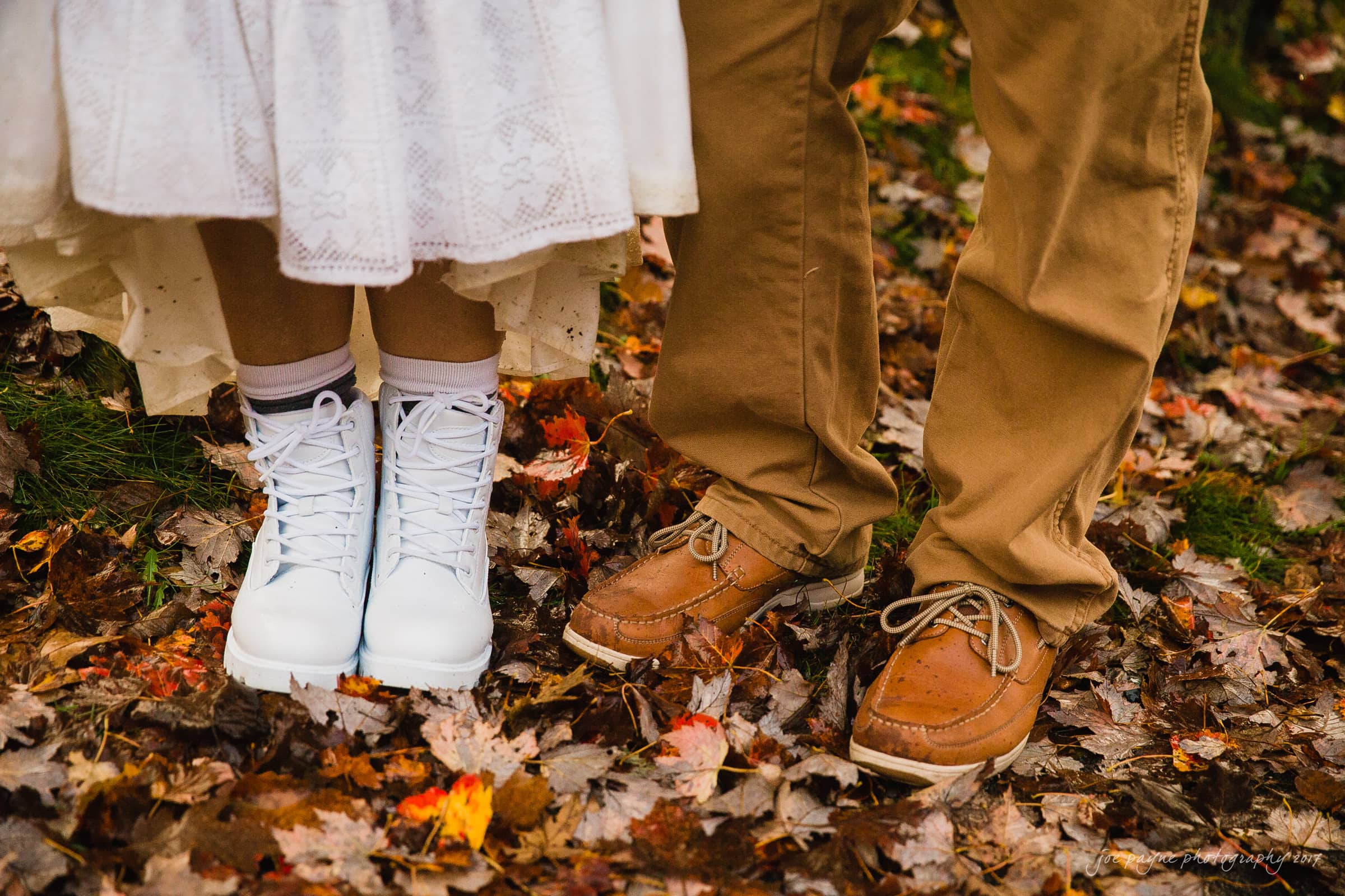 A bride in white lace sneakers and a groom in brown leather shoes stand on a carpet of autumn leaves. The brides dress is white and lacy, while the groom wears beige pants. The ground is covered in colorful fallen leaves.