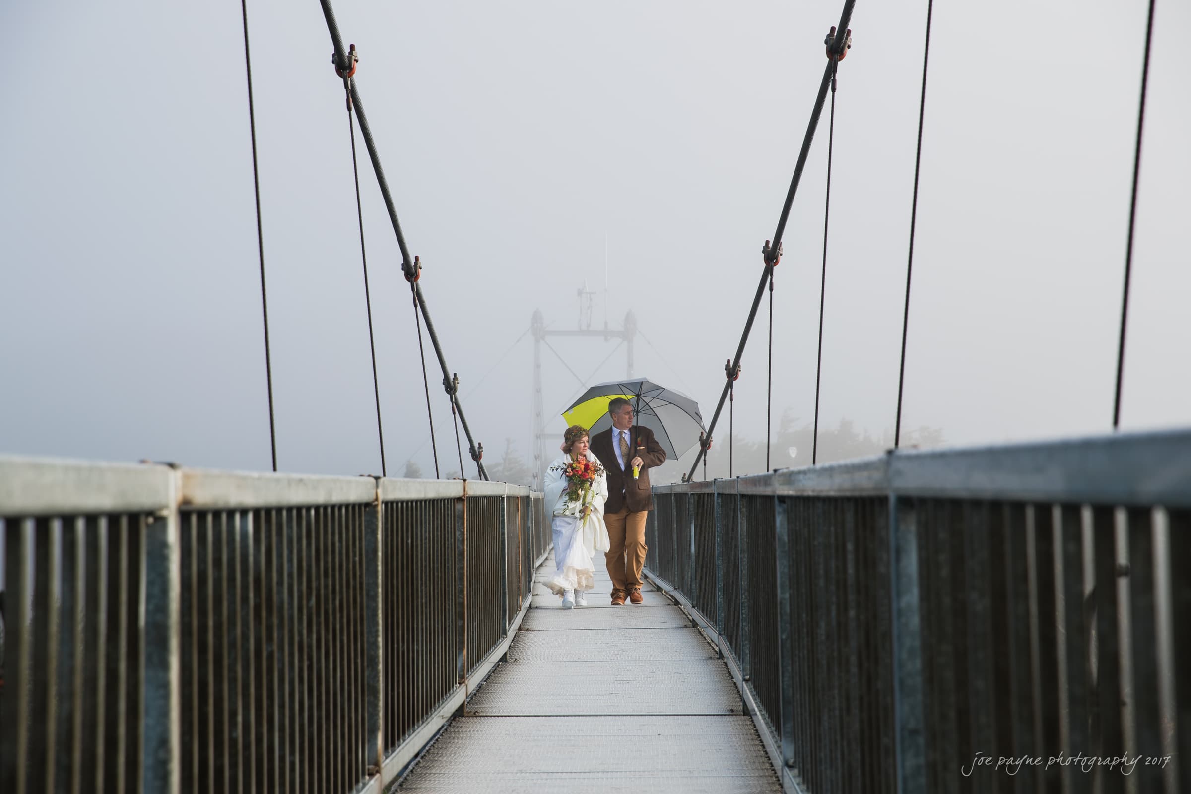 A bride and groom walk across a foggy suspension bridge. The groom holds a yellow umbrella and both are dressed in wedding attire, with the bride carrying a bouquet of flowers. The scene is enveloped in mist, creating a dreamy atmosphere.