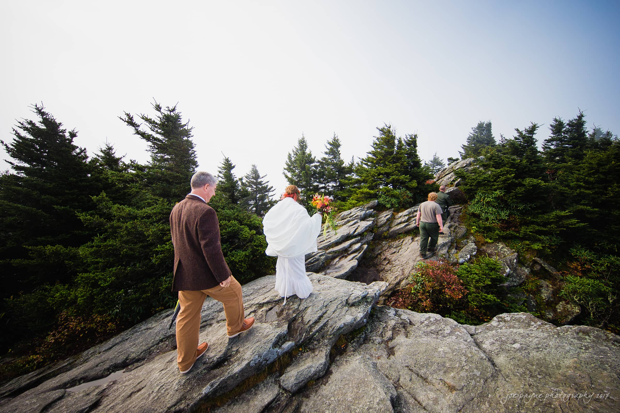 A bride in a white dress holding a bouquet walks along a rocky mountain path, accompanied by two people, one in a brown jacket and the other in casual attire. Evergreen trees surround them under a clear sky.