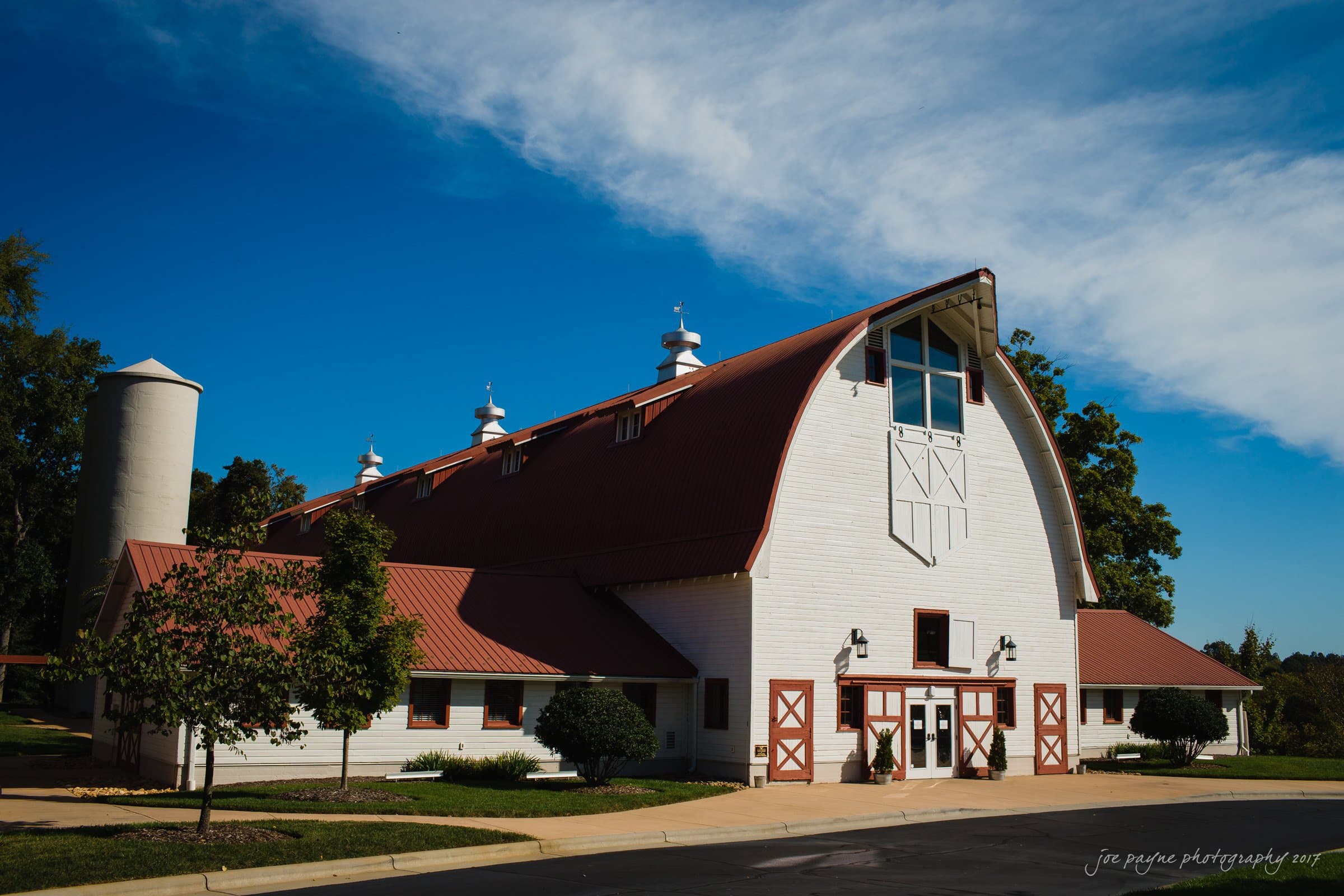 winmock nc wedding photographer - hallie & cullen A large, white barn with a red roof and cupolas stands under a bright blue sky. A cylindrical silo is adjacent on the left. The barn features multiple windows and a central entrance with double doors. Trees and a clear sky complete the scene.