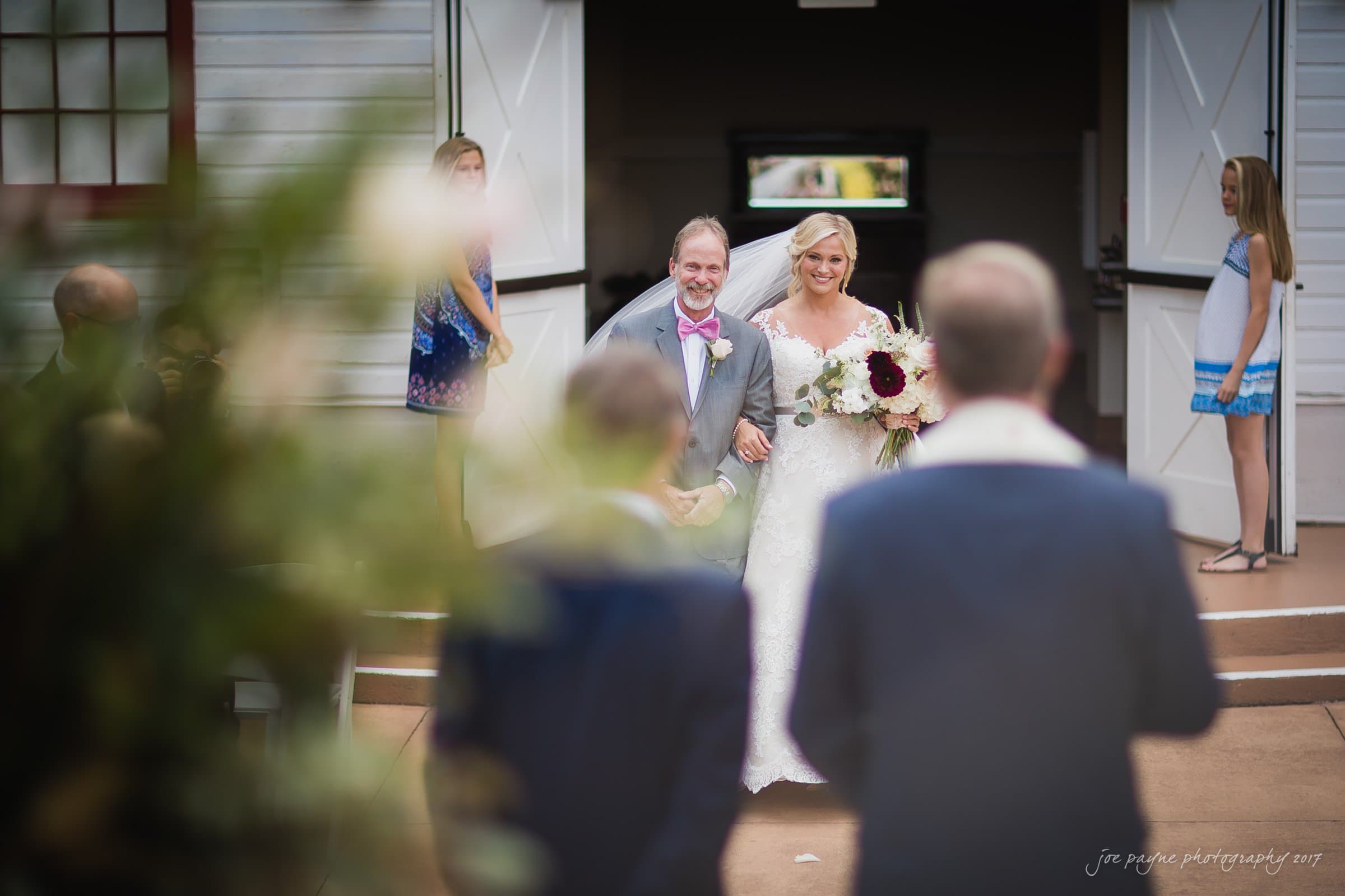 winmock nc wedding photographer - hallie & cullen A bride in a white dress walks arm in arm with a man in a suit down an aisle, surrounded by blurred greenery and guests. The pair smile warmly as they exit a building with open doors.