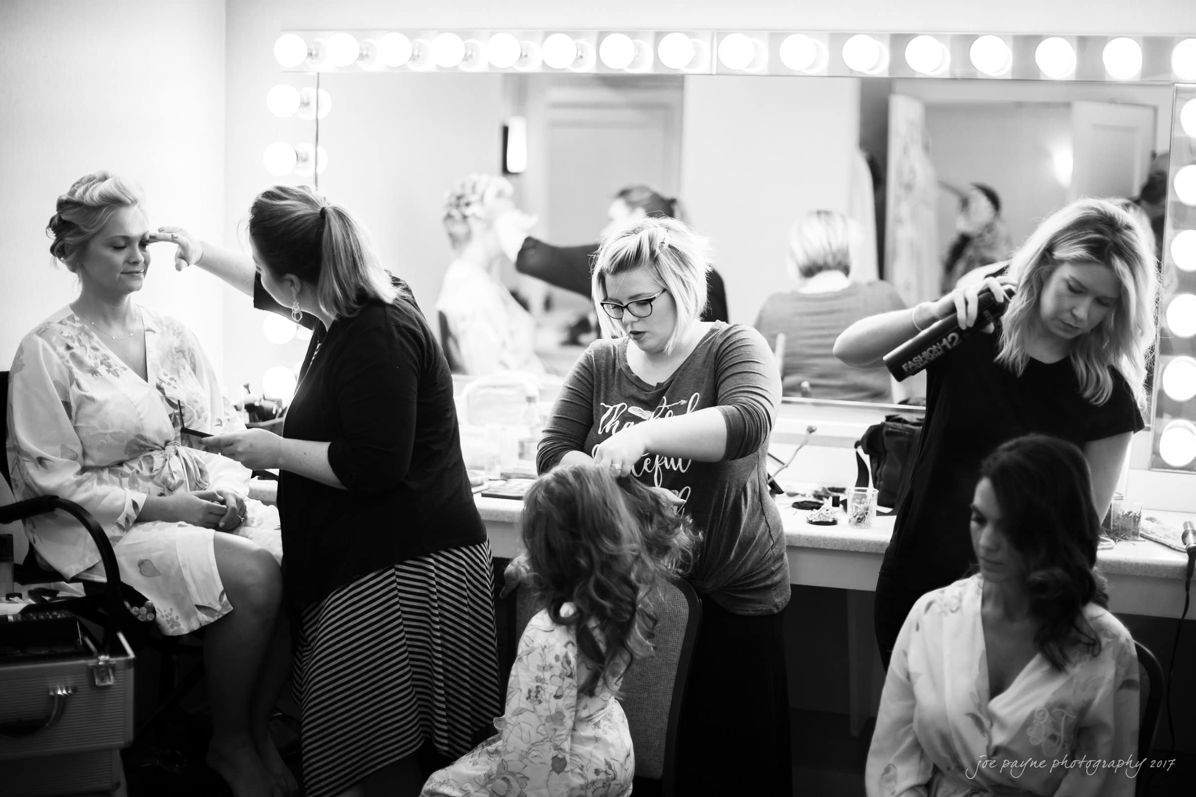 winmock nc wedding photographer - hallie & cullen A black and white photo showing a busy dressing room with three women getting ready. Stylists are working on their hair and makeup. Vanity mirrors with lights frame the background, reflecting some of the activity.