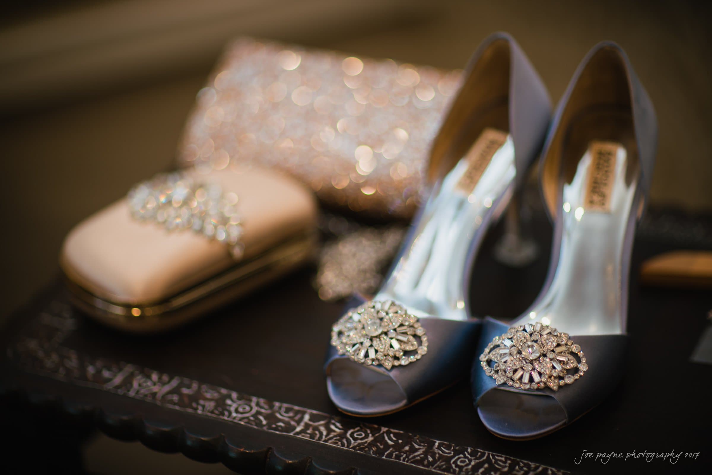 winmock nc wedding photographer - hallie & cullen Elegant pair of open-toed high heels with jeweled embellishments displayed on a dark wooden table. A sparkling clutch with similar jeweled details is placed nearby, creating a sophisticated and glamorous setting.