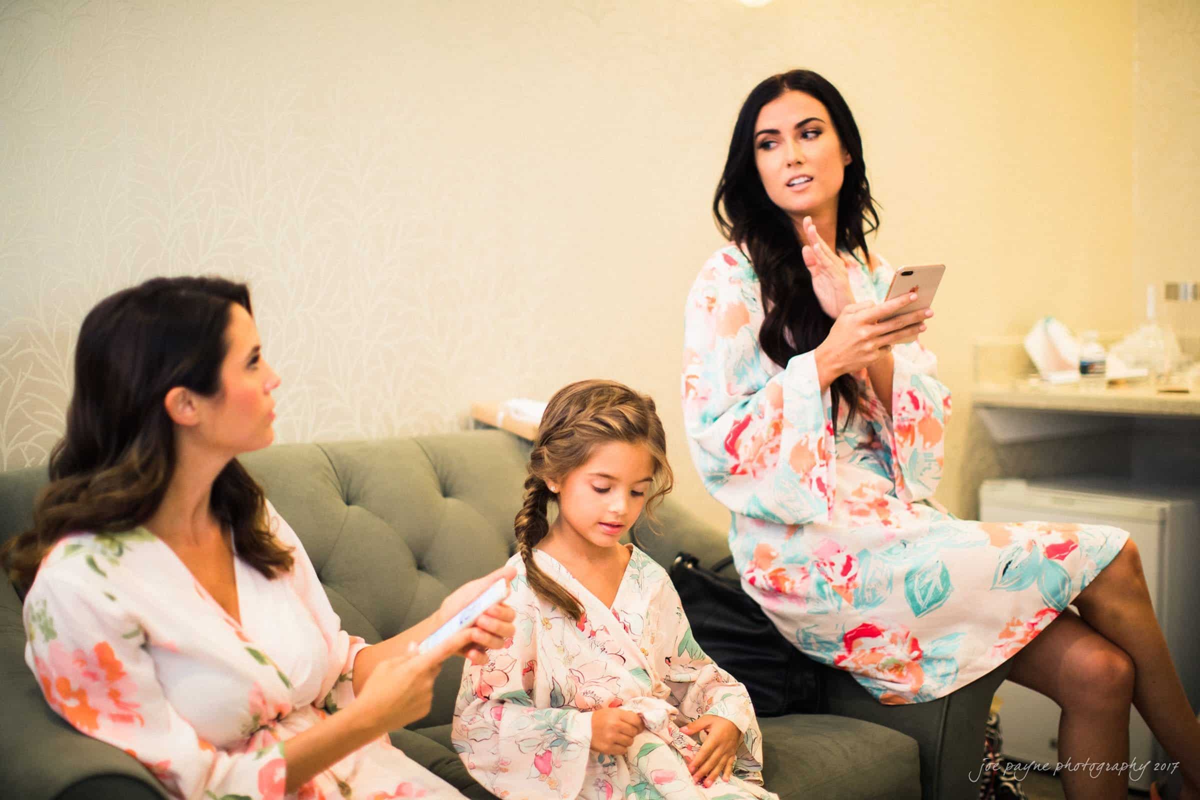winmock nc wedding photographer - hallie & cullen Three women in floral robes sit on a couch indoors, engaged in conversation. A young girl with braided hair joins them, also in a matching robe. One woman holds a smartphone, gesturing as she talks. The setting is relaxed and intimate.