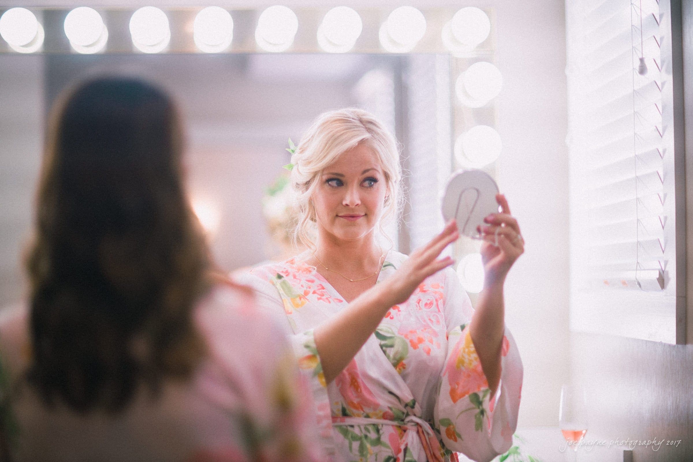 winmock nc wedding photographer - hallie & cullen A woman in a floral robe stands in front of a mirror with illuminated bulbs, holding a compact mirror. She is focused on her reflection, with a soft background and a glass of wine on the right.