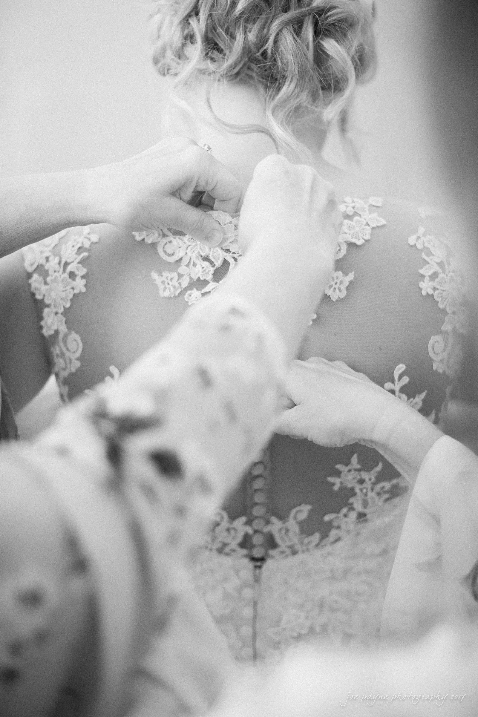winmock nc wedding photographer - hallie & cullen A bride in a lace wedding dress is being helped with buttoning up the back of her dress. Two people are assisting her, their hands visible and focused on the intricate lace details. The image is in black and white.