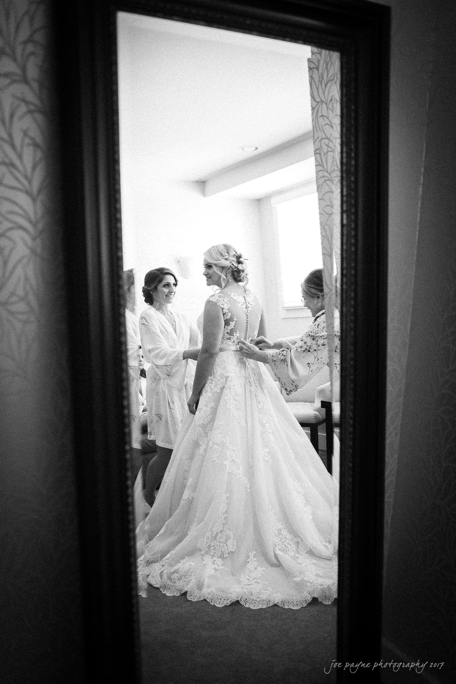 winmock nc wedding photographer - hallie & cullen A bride in an elegant lace gown is getting ready with the help of two women in a softly lit room. The scene is reflected in a large mirror, capturing the bridal preparations and camaraderie.