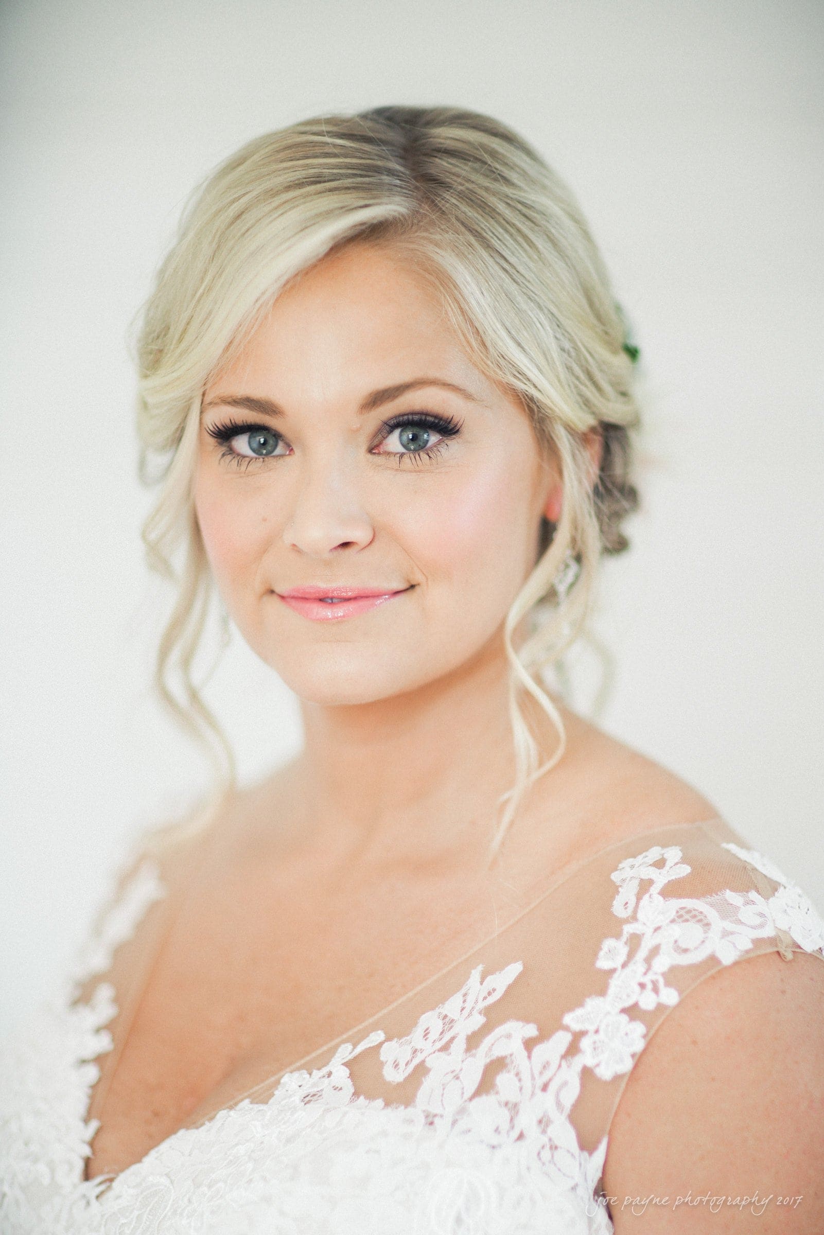 winmock nc wedding photographer - hallie & cullen A woman with styled blonde hair and blue eyes smiles softly. She wears a lace-embellished dress, suitable for a wedding. The background is softly blurred, focusing on her serene and elegant expression.