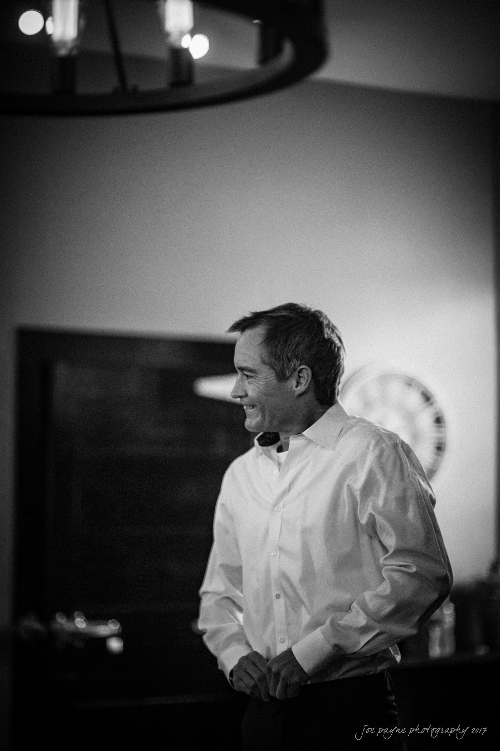 winmock nc wedding photographer - hallie & cullen A man in a white dress shirt is smiling and standing indoors. The background includes a clock and a doorway. The image is in black and white, with soft lighting creating a warm ambiance.