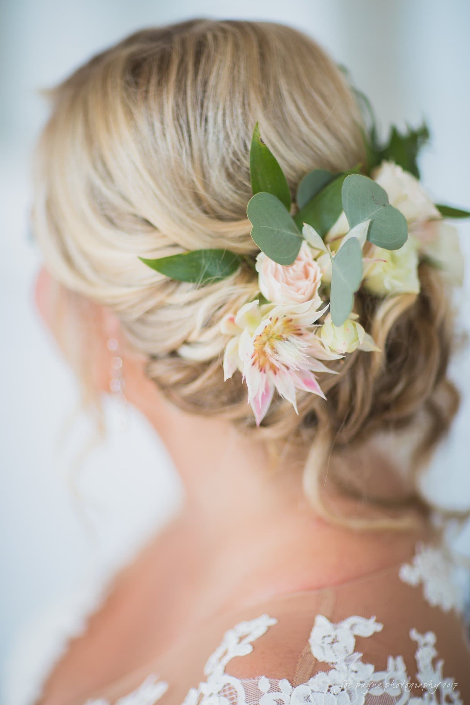 winmock nc wedding photographer - hallie & cullen A bride with blonde hair styled in an elegant updo, adorned with pastel flowers and green leaves. She is wearing lace-trimmed wedding attire, with soft-focus detailing emphasizing the floral accents.