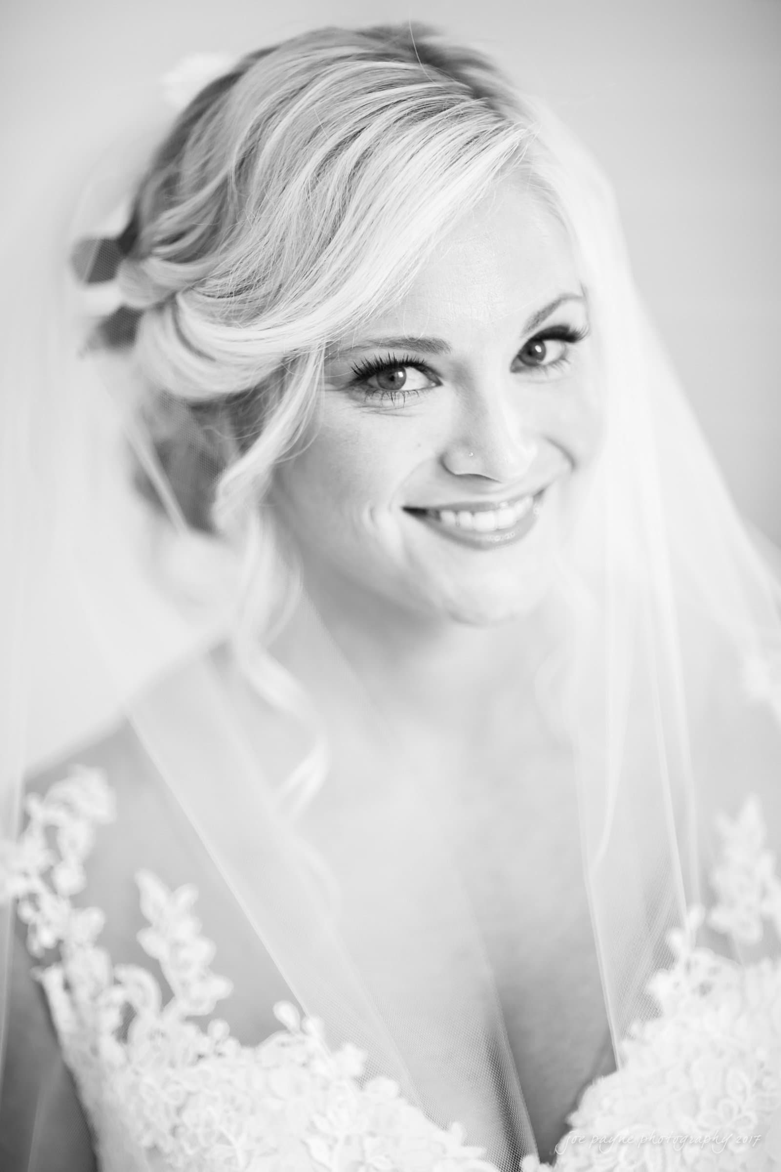 winmock nc wedding photographer - hallie & cullen A black and white portrait of a bride with blonde hair, smiling warmly. She is wearing a lace wedding dress and a veil, and her hair is styled elegantly with soft curls.