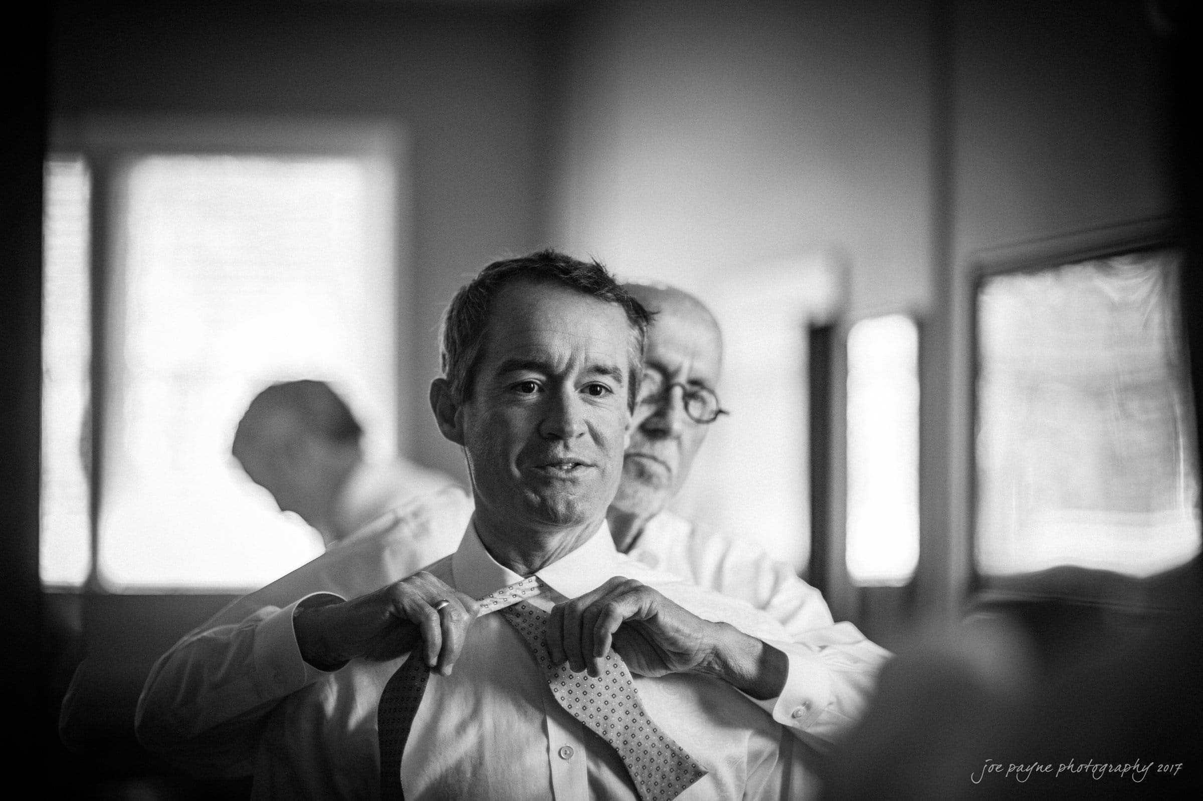 winmock nc wedding photographer - hallie & cullen Man in a suit being helped with his tie by another man in a room. The background features blurred figures and window light, creating a warm atmosphere. Black and white photograph.