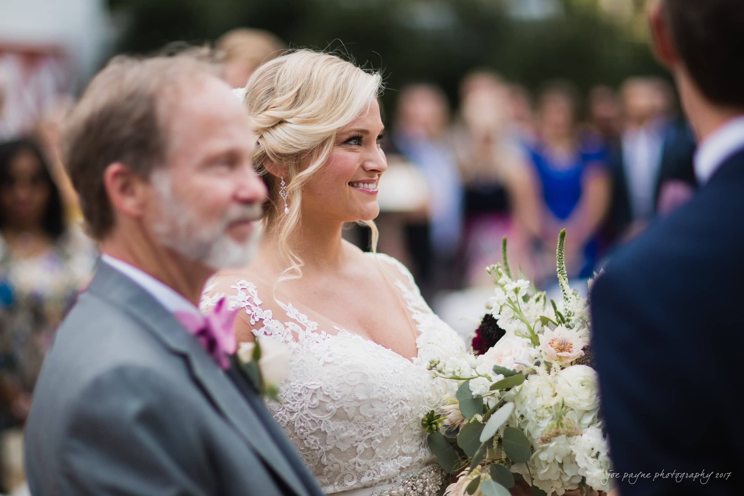 winmock nc wedding photographer - hallie & cullen A bride in a white lace dress holds a bouquet and smiles, standing next to an older man in a gray suit with a pink boutonniere. A crowd in blurred background watches the couple.