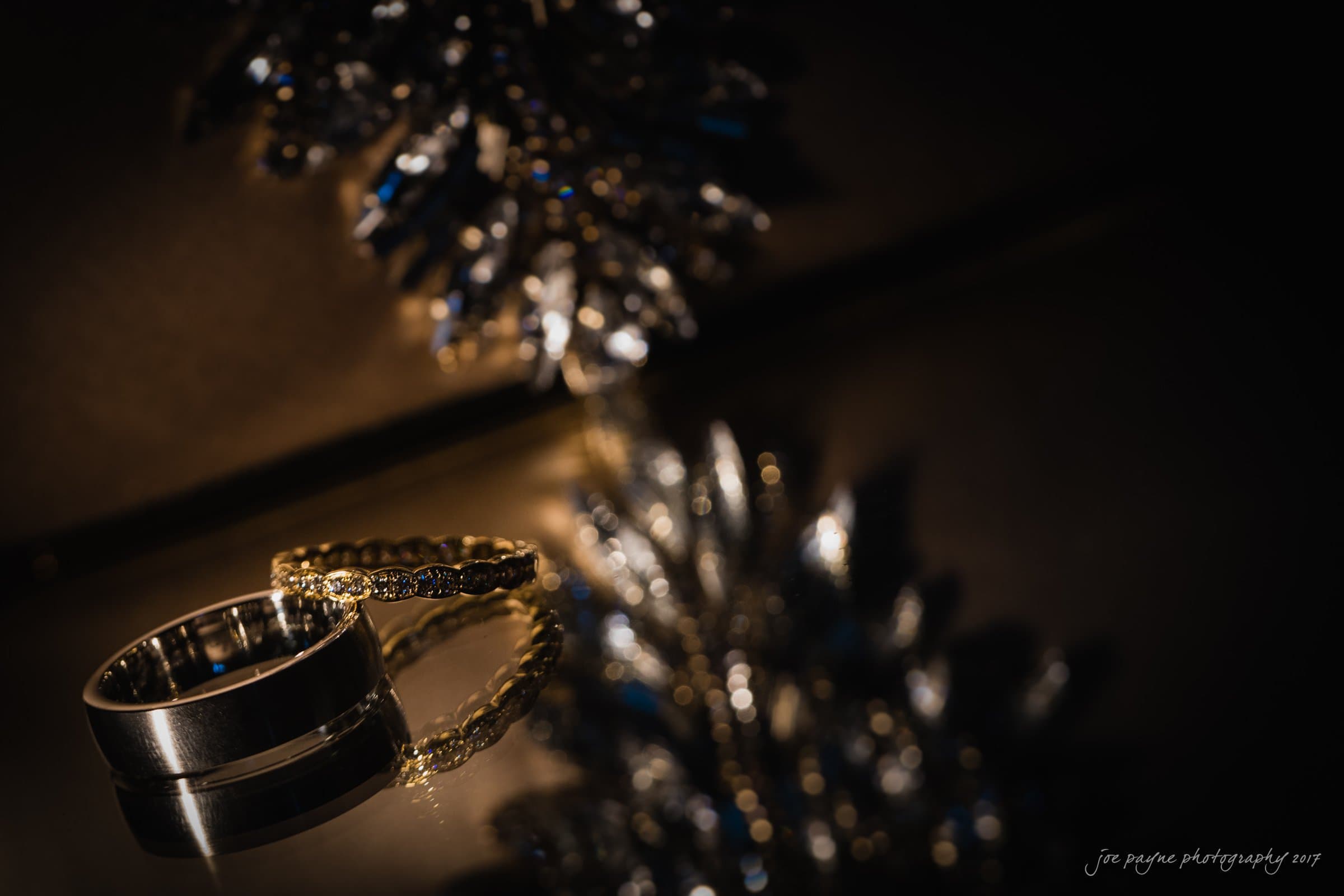 winmock nc wedding photographer - hallie & cullen A close-up shot of two elegant rings: one gold with small diamonds, and one plain silver. They rest on a reflective surface, capturing light from a nearby luminous, decorative object with a starburst design.