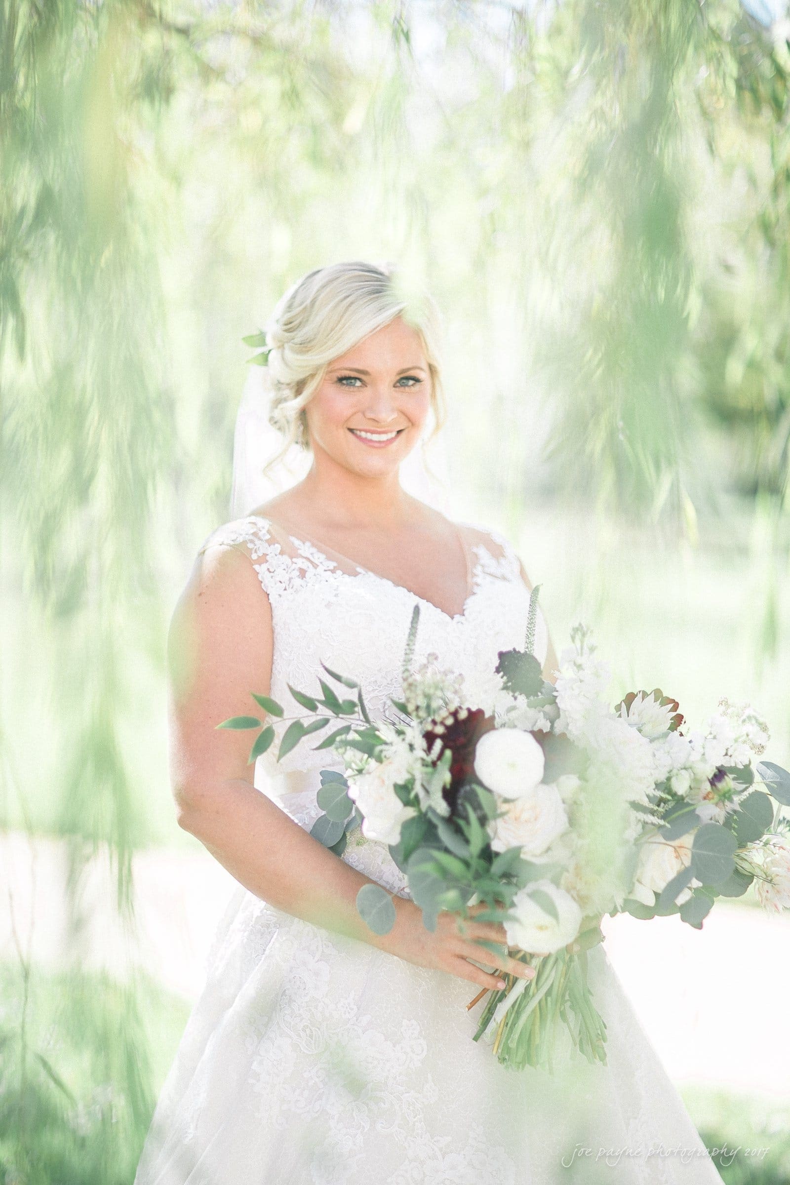 winmock nc wedding photographer - hallie & cullen A bride in a white lace wedding dress holds a bouquet of white and dark flowers and greenery. She stands under a canopy of soft, blurred green leaves, smiling at the camera, with sunlight filtering through the foliage.