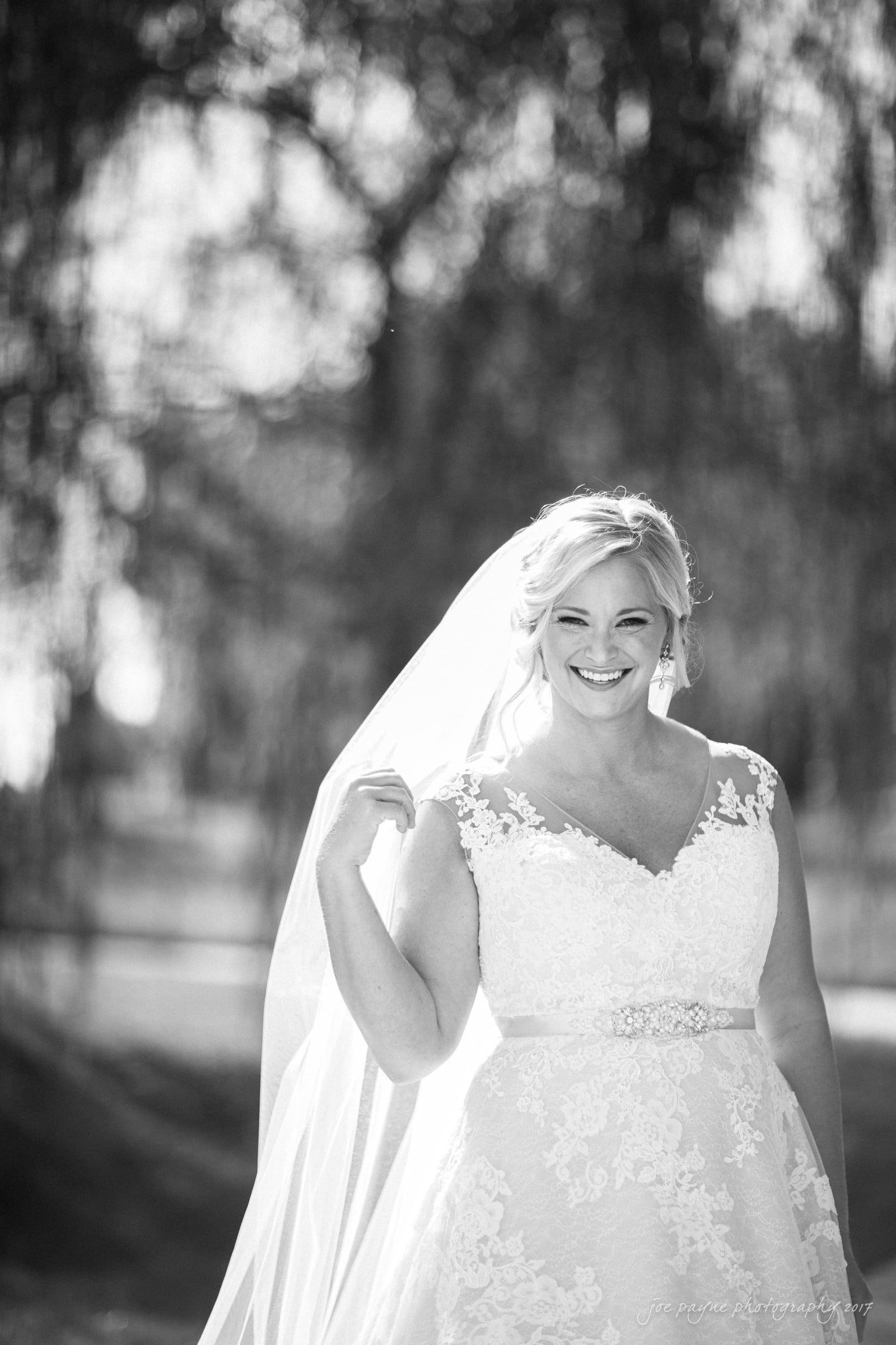 winmock nc wedding photographer - hallie & cullen A bride in a lace wedding dress smiles while holding her veil. She is standing outdoors with blurred trees in the background, captured in black and white.