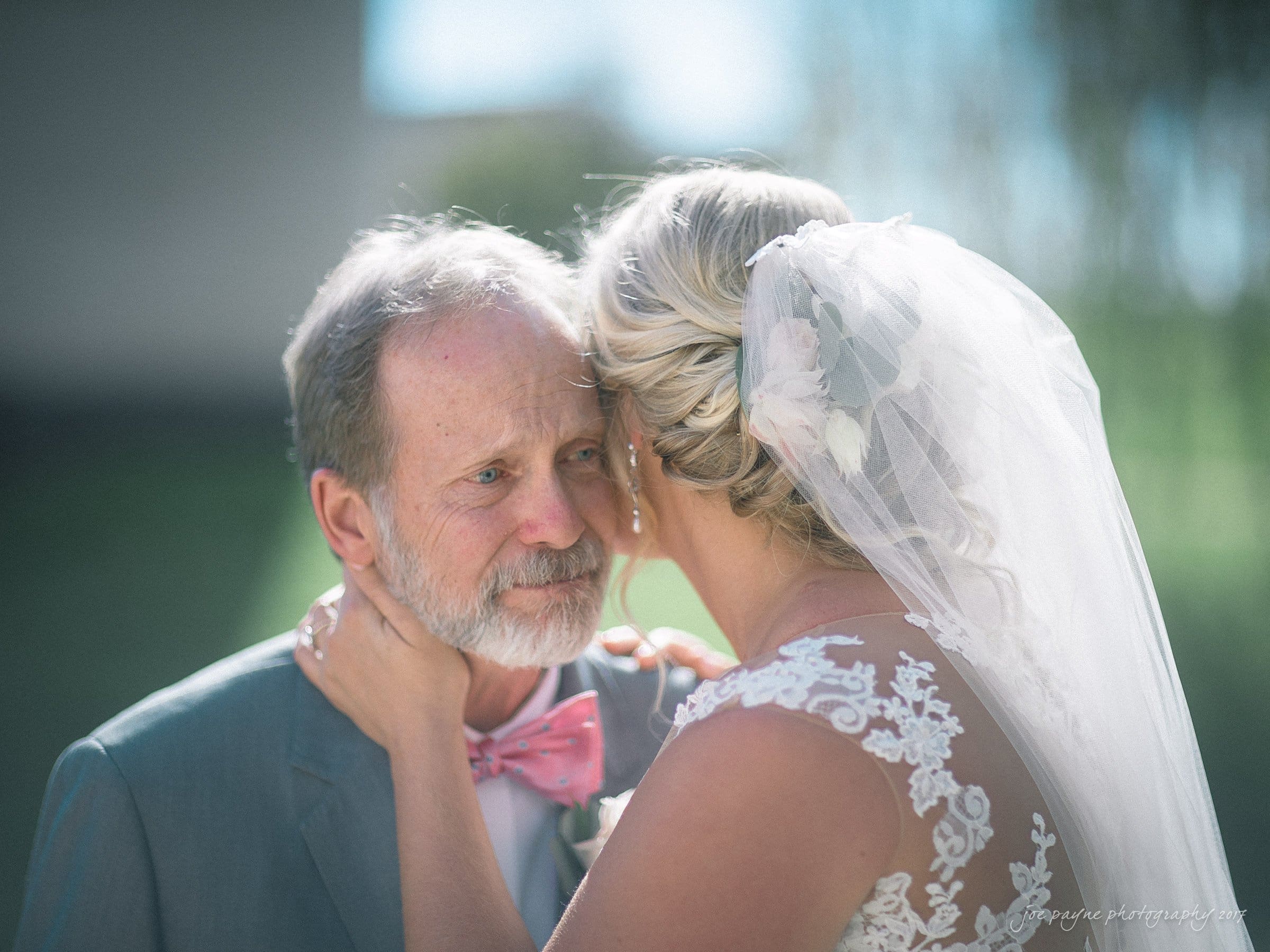 winmock nc wedding photographer - hallie & cullen A bride with a lace veil holds an older mans face tenderly. The man wears a gray suit with a pink bow tie. They are outdoors with soft-focus greenery in the background, sharing a heartfelt, intimate moment.