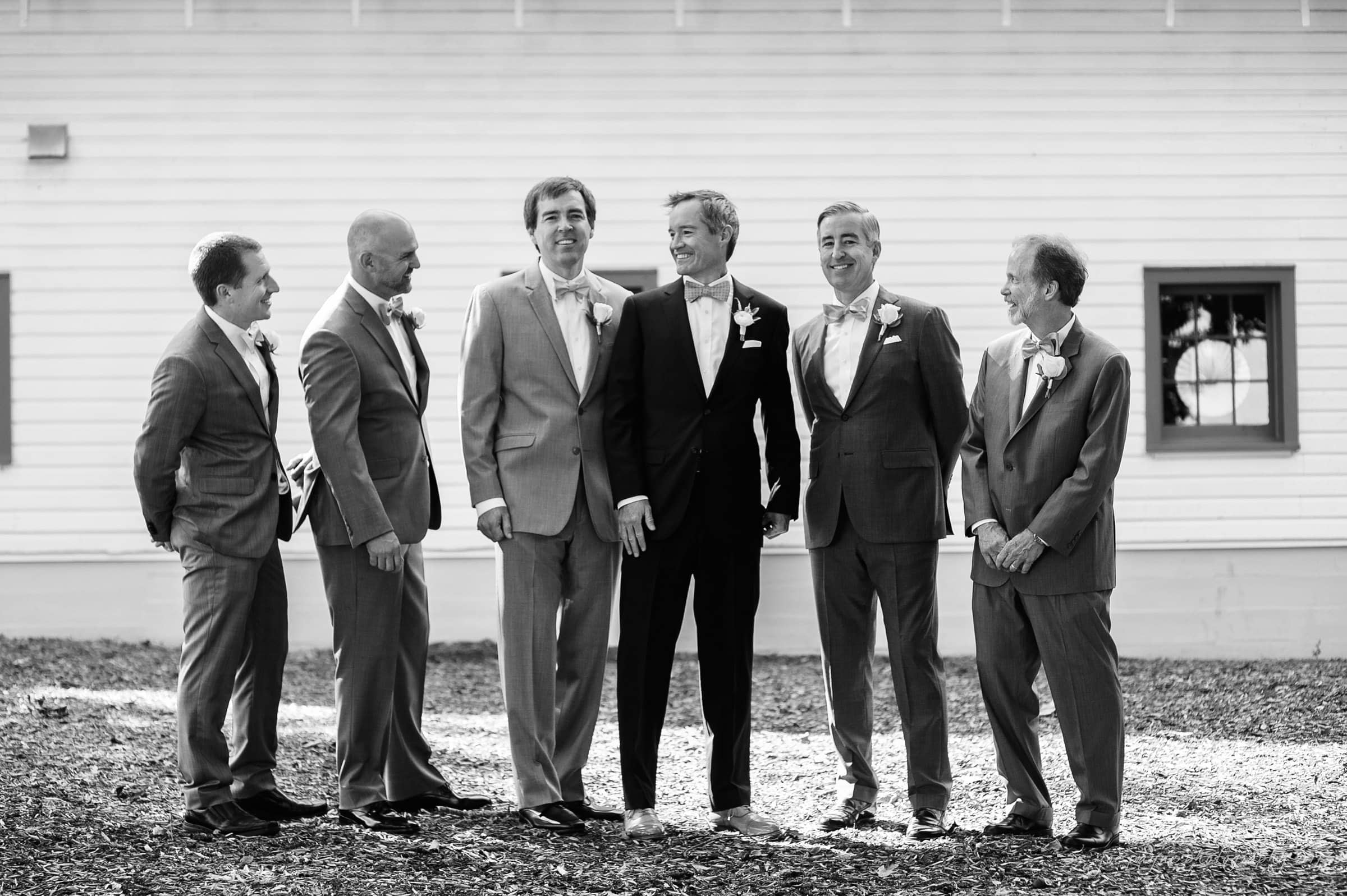 winmock nc wedding photographer - hallie & cullen A group of six men in suits stand outside in front of a white building, smiling and talking. Five men wear light-colored suits, and one man in the center wears a dark suit. The setting appears formal, possibly at a wedding. Black and white photo.